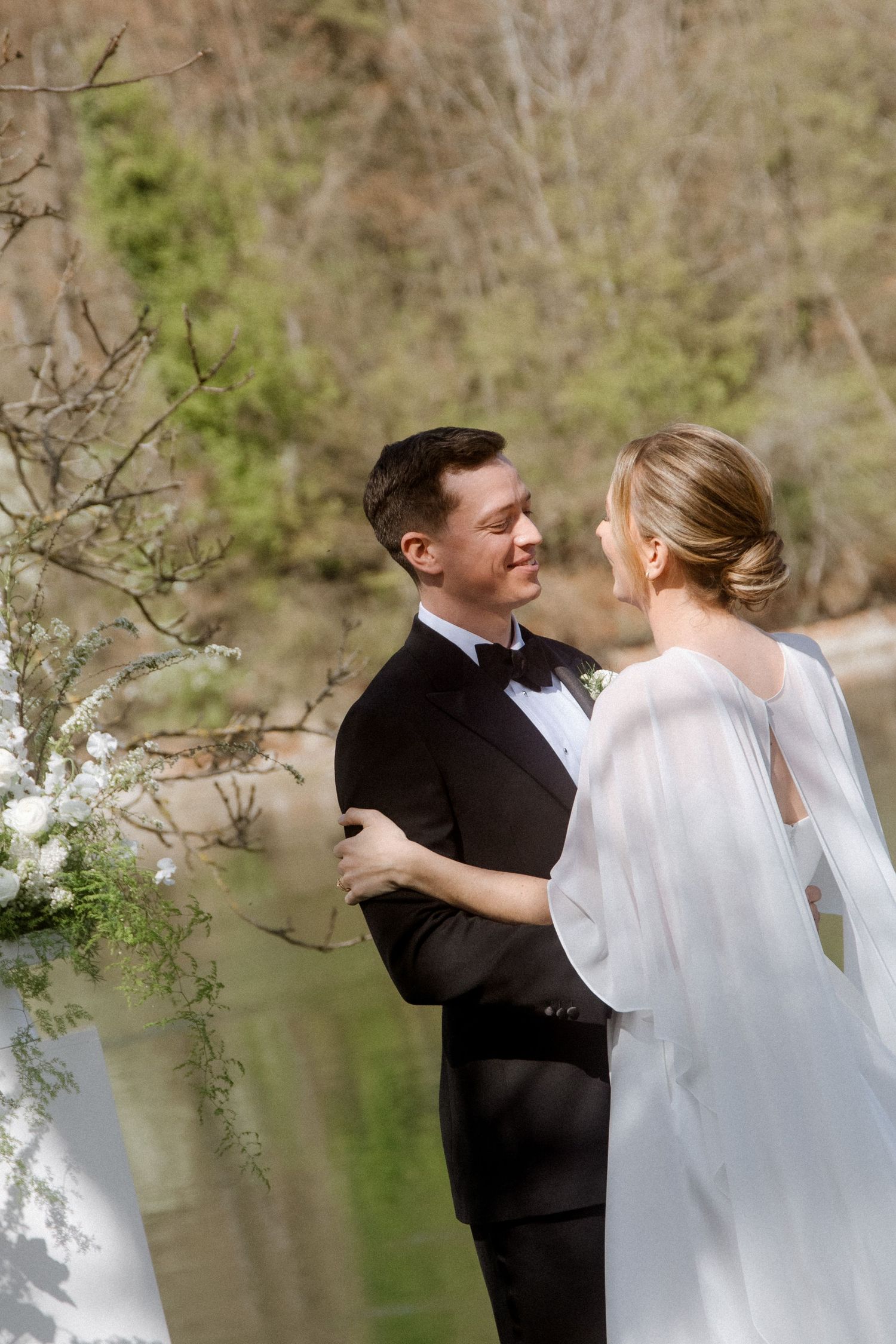 Smiling groom in black tuxedo embraces bride in white cape dress during outdoor lakeside wedding ceremony.