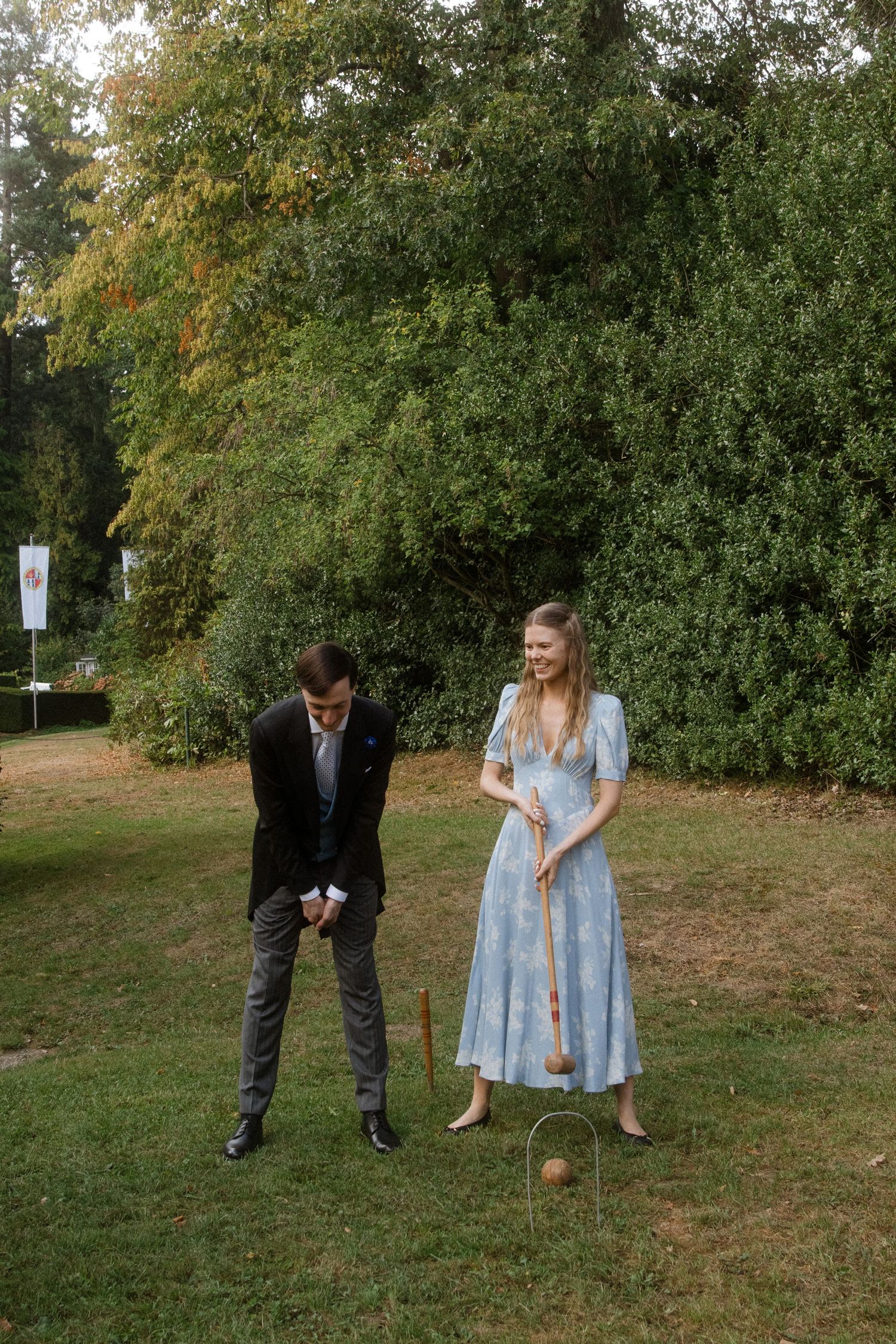 Two people playing croquet on a grassy lawn surrounded by trees.