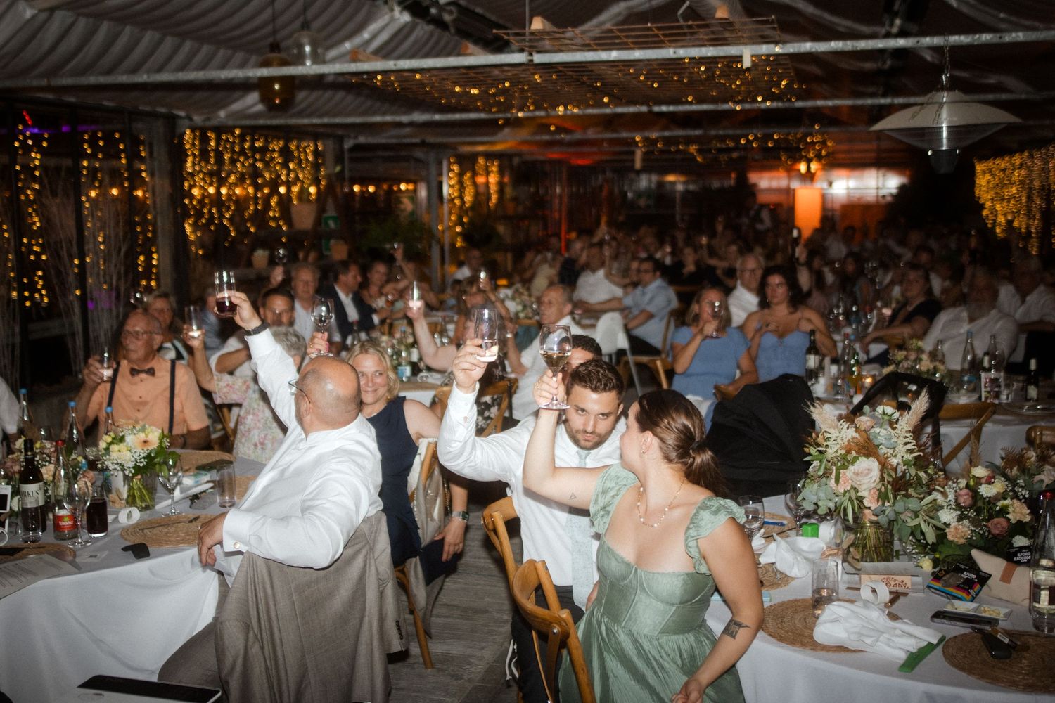Wedding reception guests celebrating with raised glasses in a decorated venue with string lights.