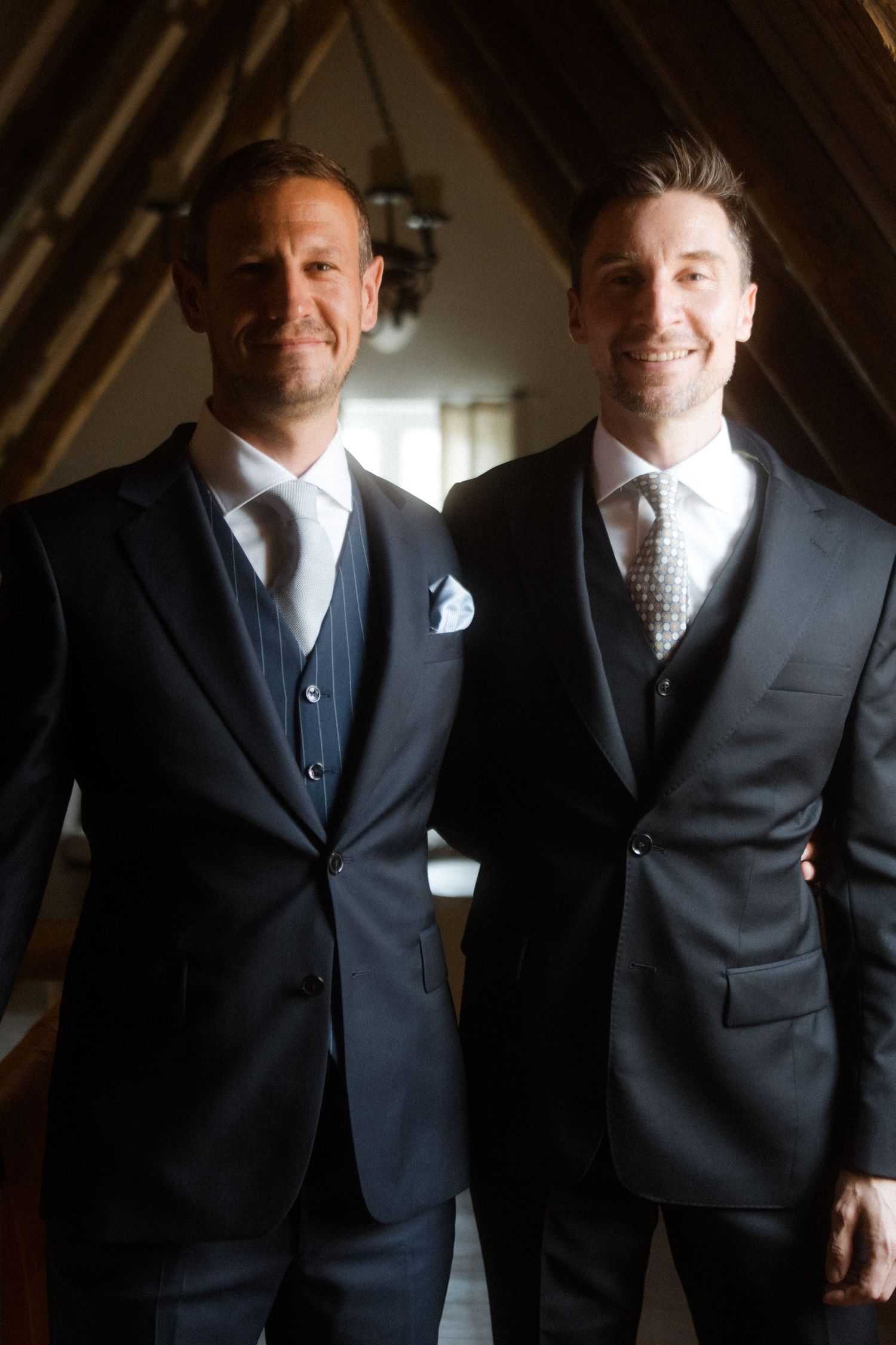 Two men in dark formal suits pose together in an attic room with wooden beams.