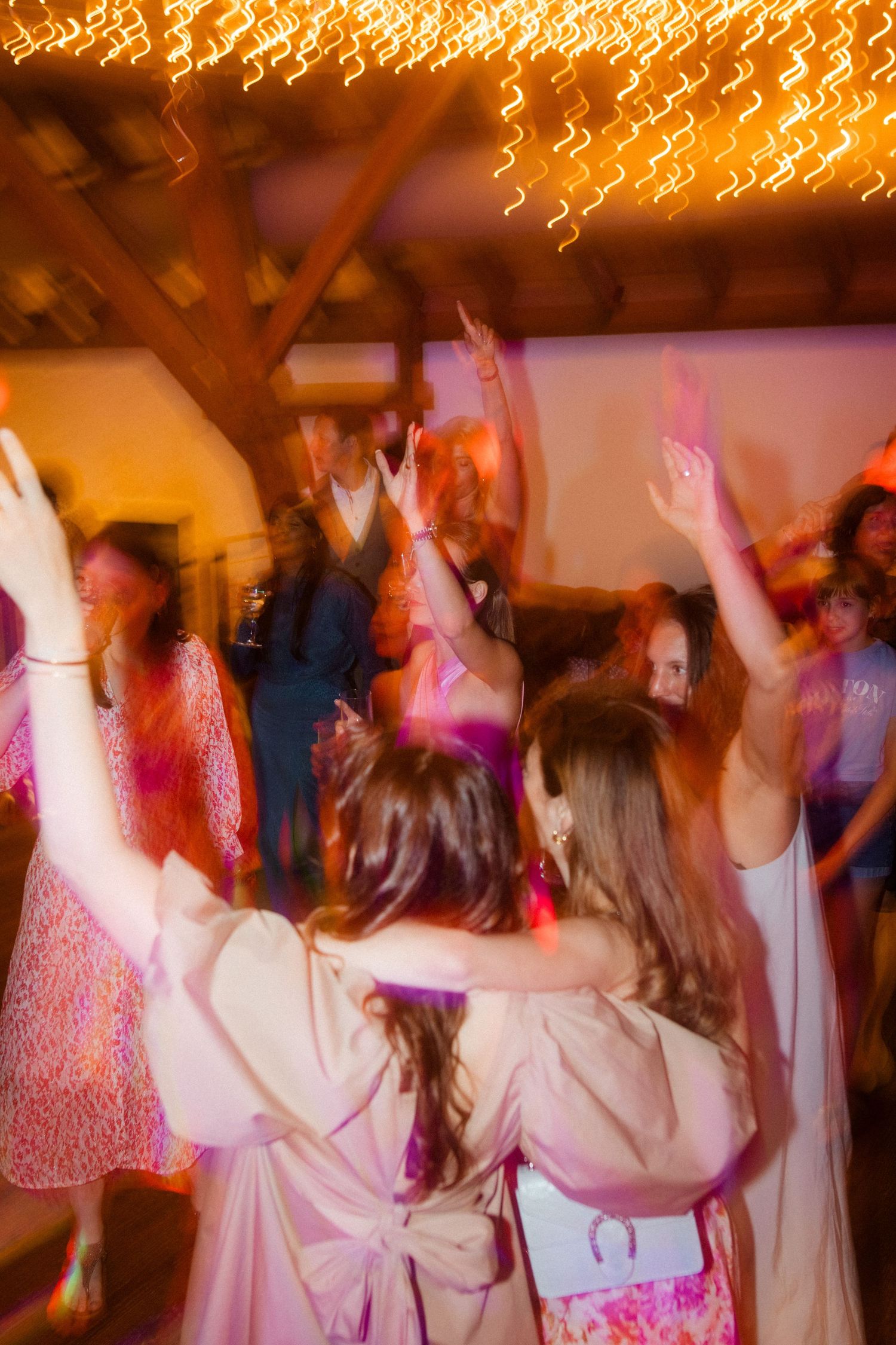Party guests dancing with raised arms under warm glowing string lights.