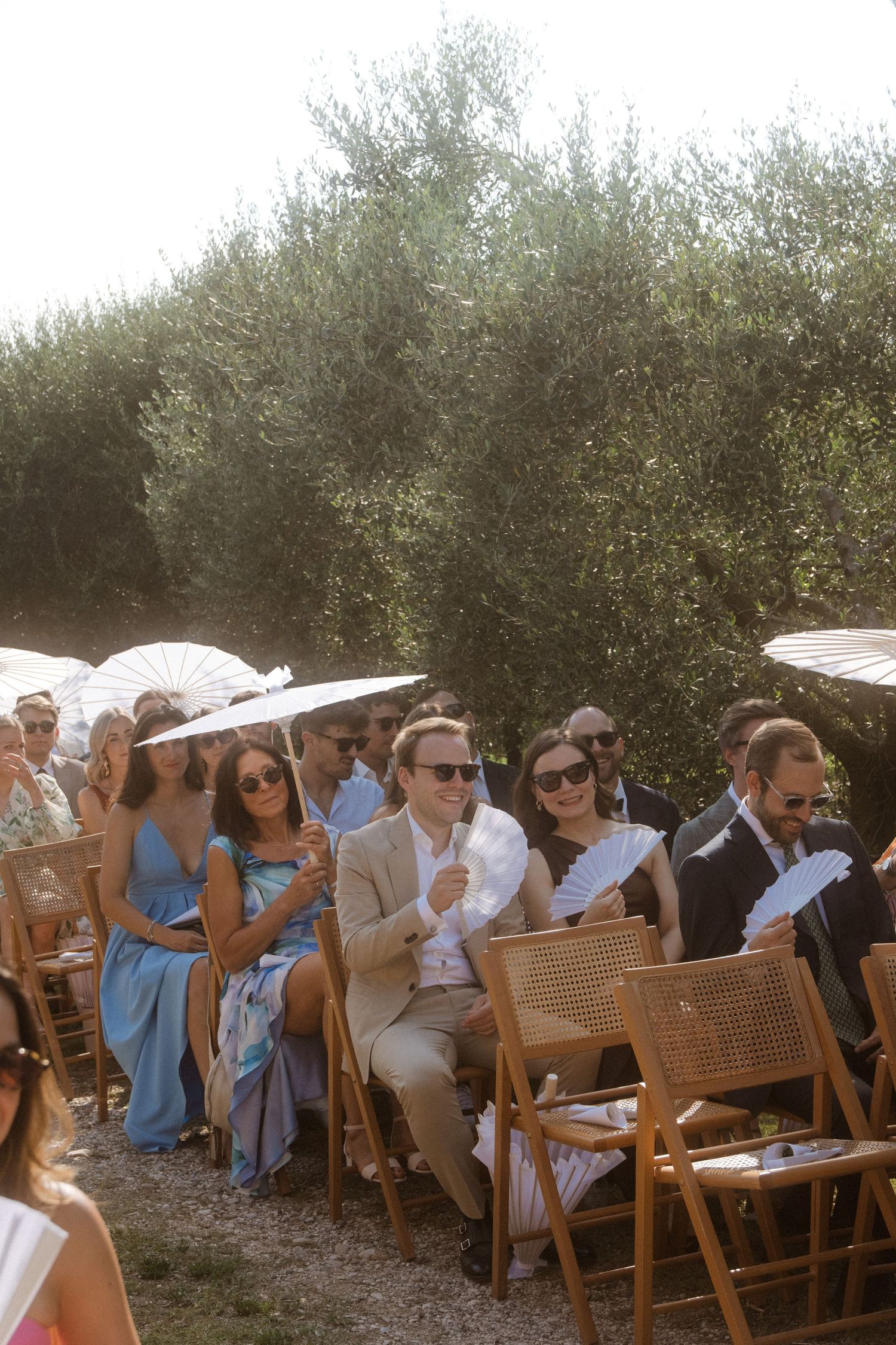 Wedding guests with parasols seated on wooden chairs during an outdoor ceremony.
