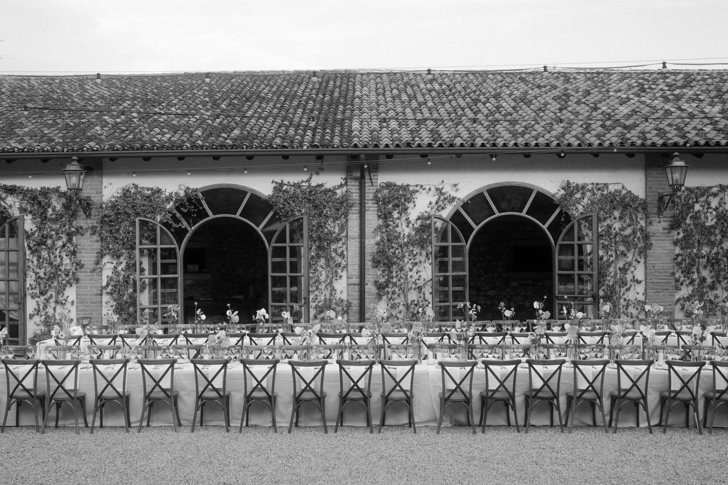 Long rustic stone building with arched windows and climbing vines in black and white.