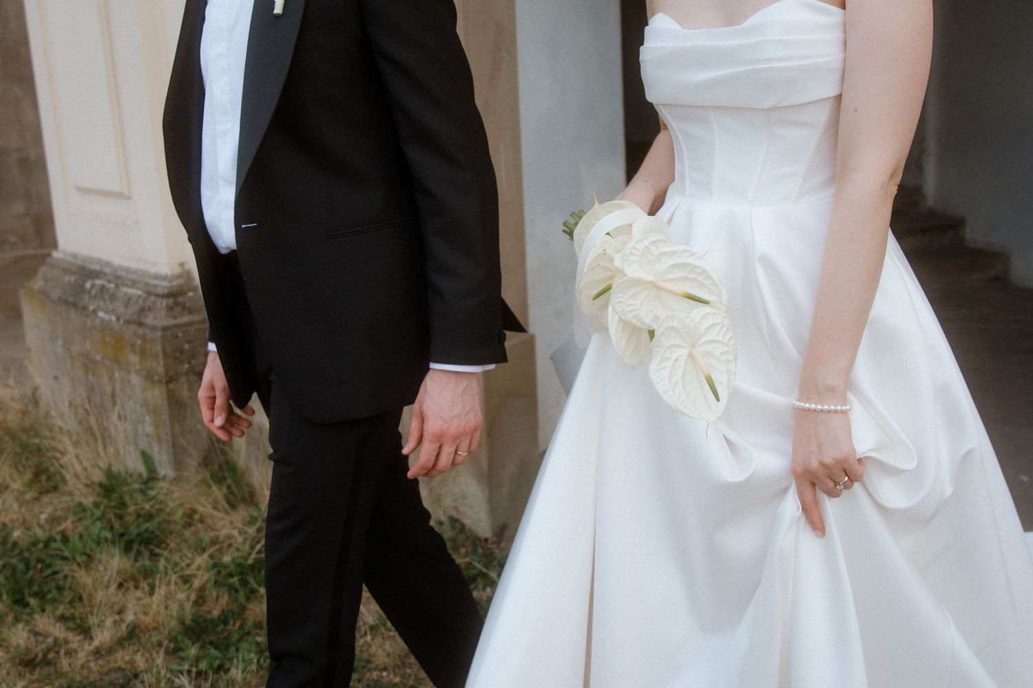 A bride and groom walk together in formal wedding attire against a stone wall backdrop.