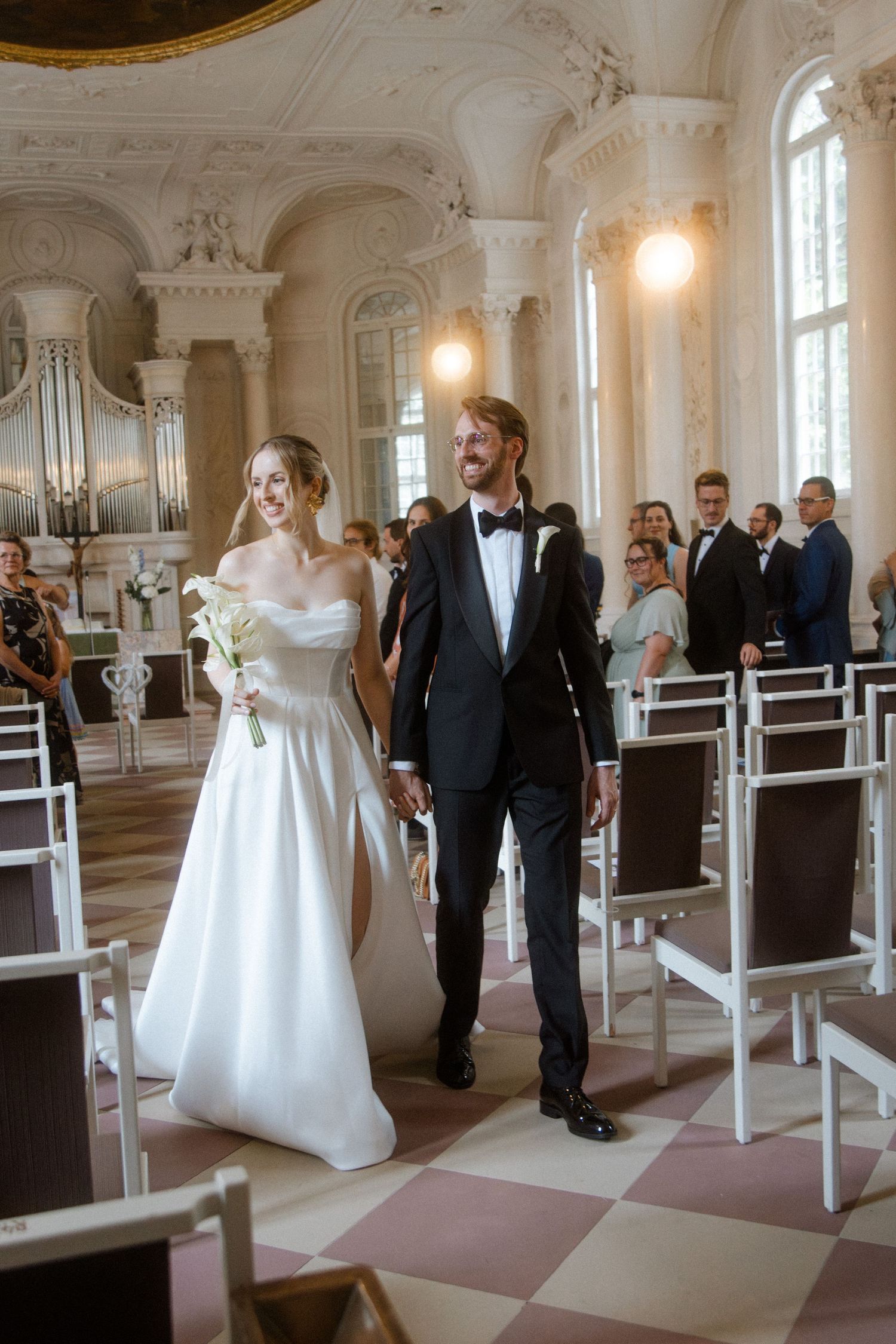 Couple walks down church aisle with ornate pipe organ and classical architecture in background.