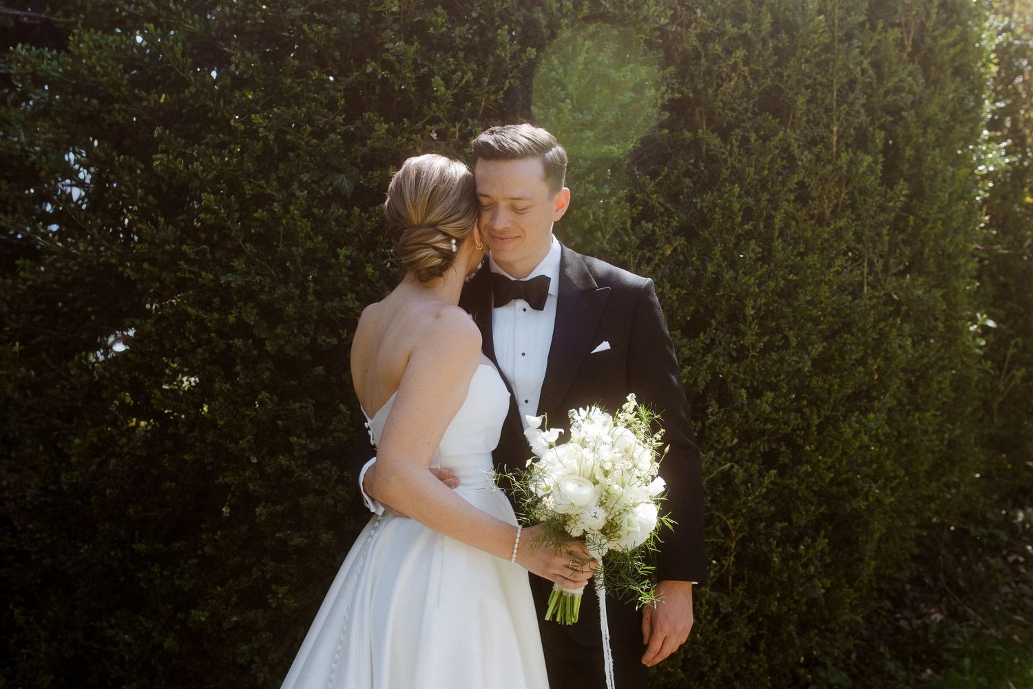 Bride and groom share a romantic moment in front of lush green hedges, bride holding white floral bouquet.