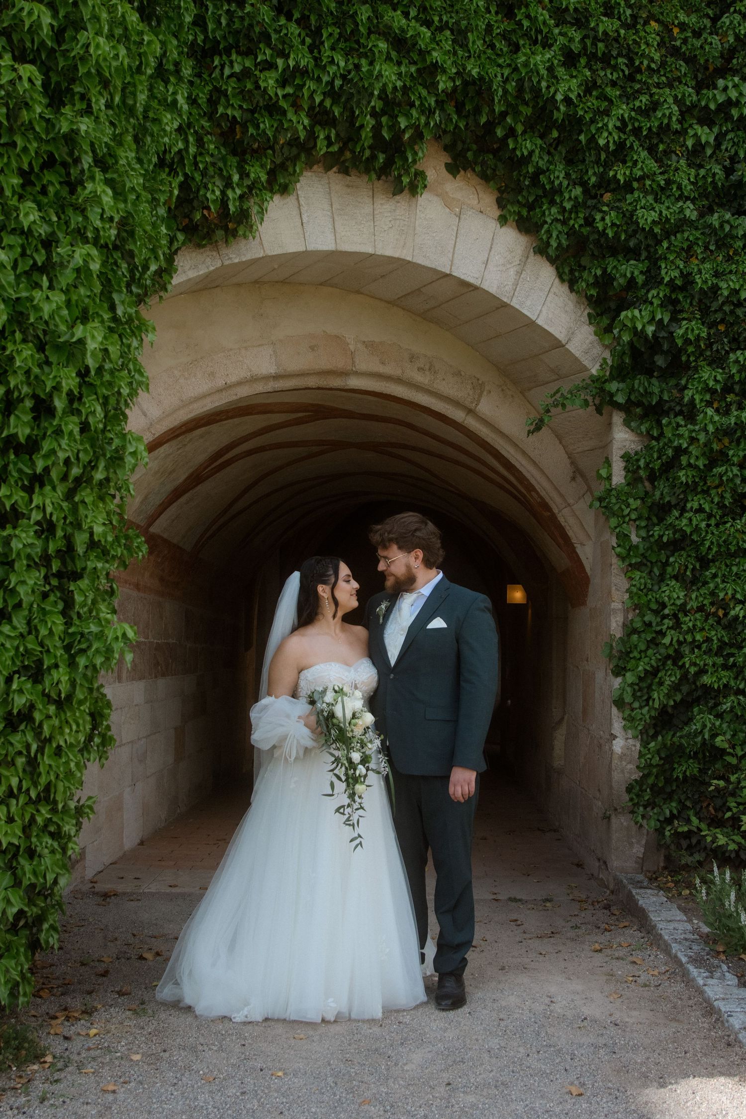 A couple in wedding attire standing in a stone archway covered with green ivy.