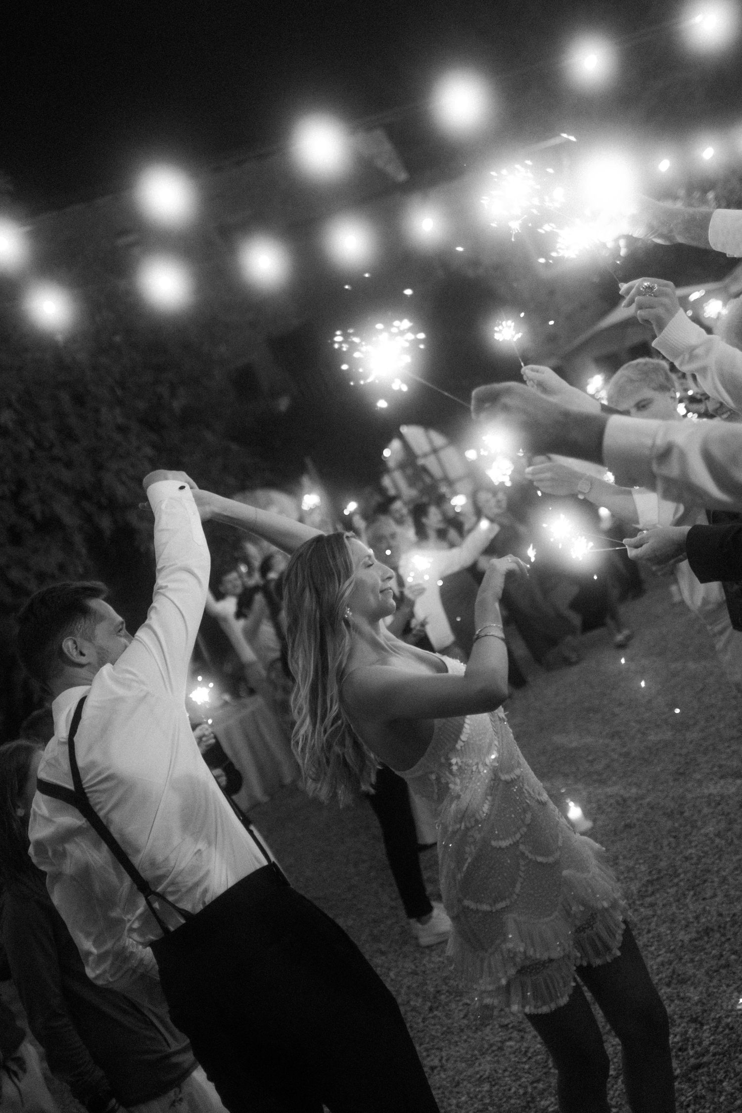 Wedding guests celebrate at night under string lights with raised glasses.