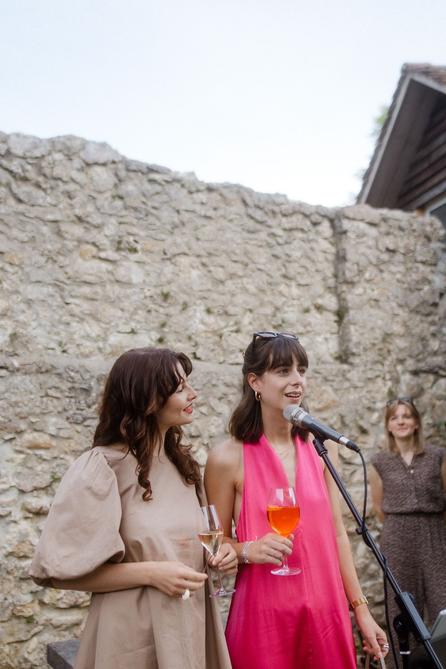 Two women in contrasting pink dresses share a laugh against a rustic stone wall.