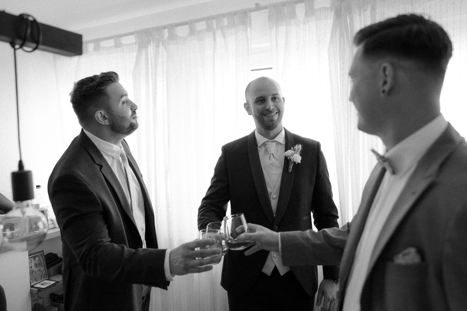 Groomsmen sharing drinks and conversation in formal attire by a window.