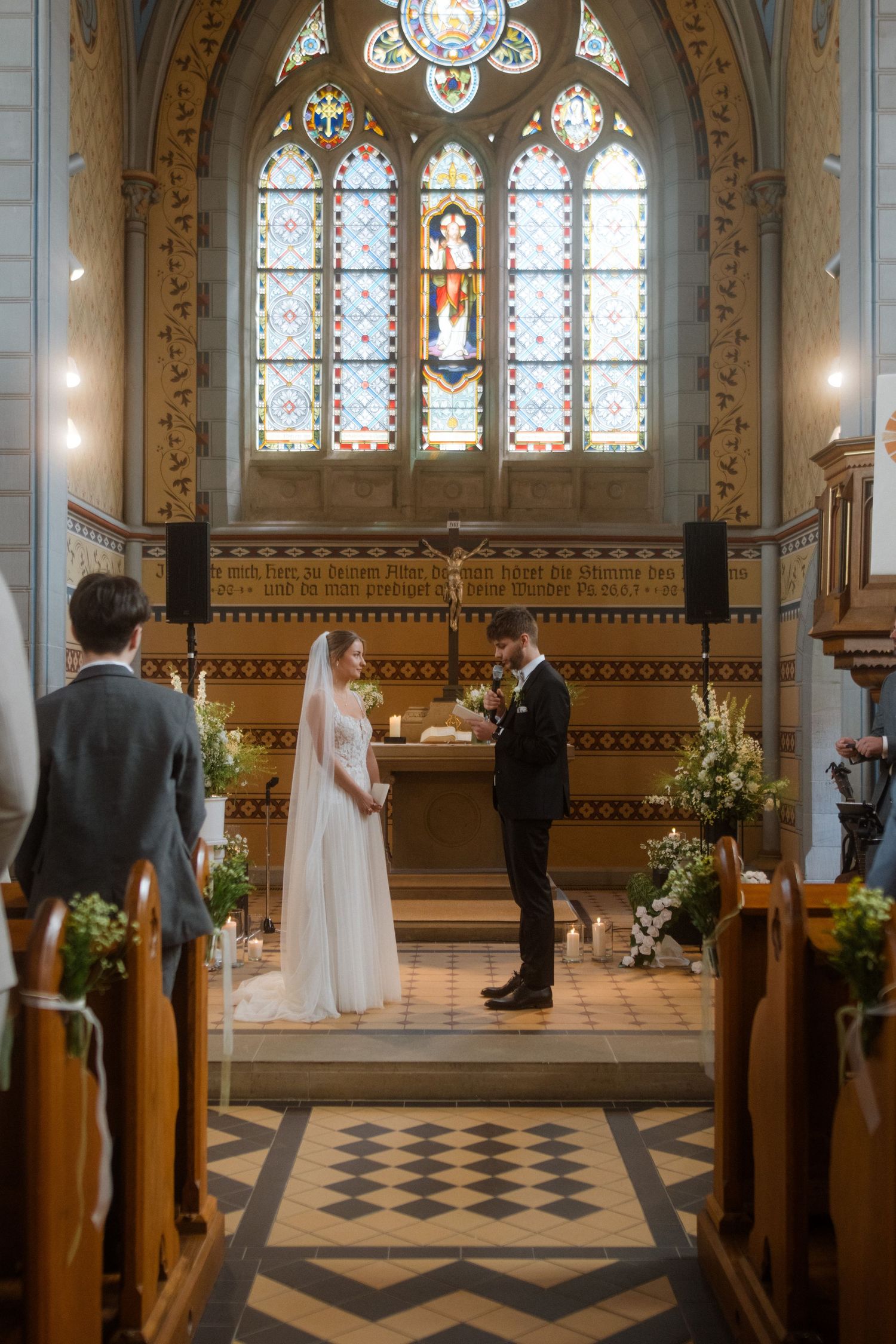 Wedding ceremony in a church with stained glass windows and checkered floor pattern.