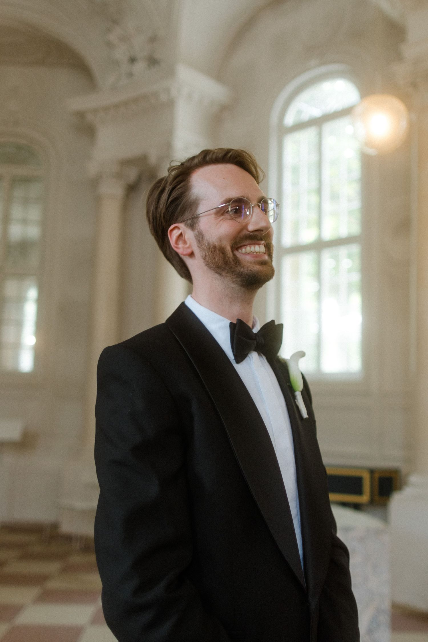 A groom in a classic black tuxedo and bow tie smiling in an ornate chapel.
