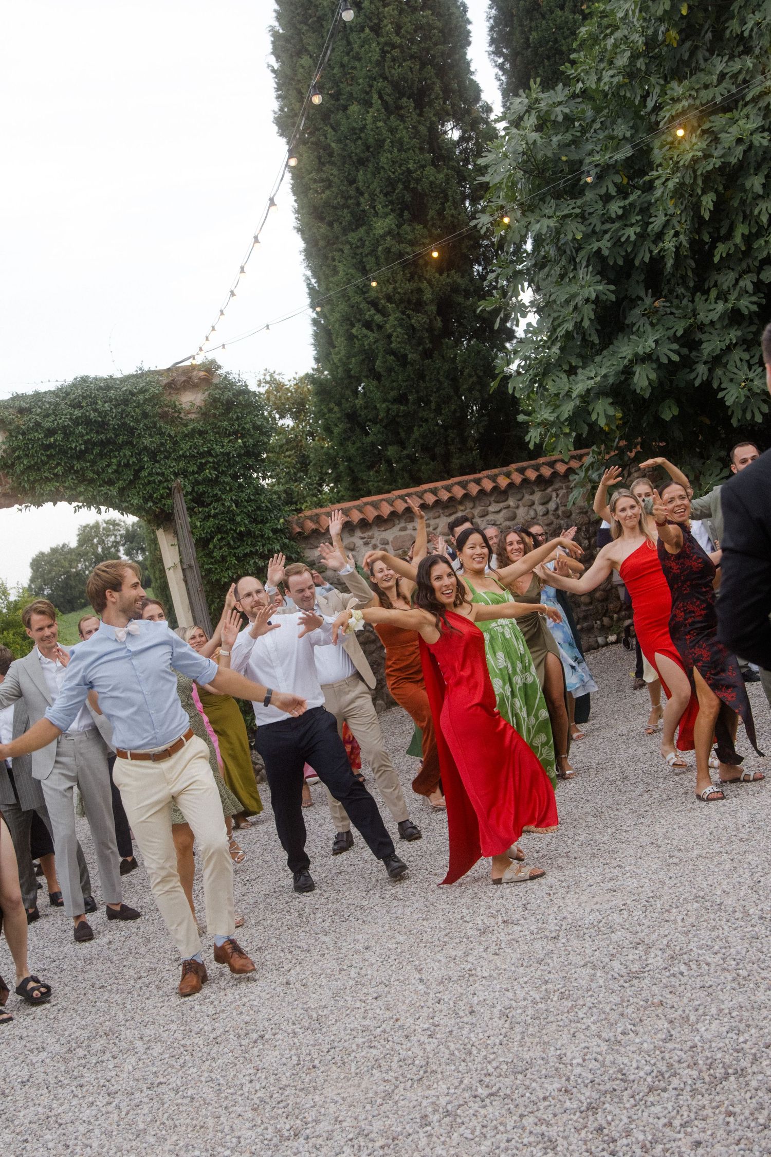 Wedding party performs choreographed dance moves in colorful attire on gravel patio.