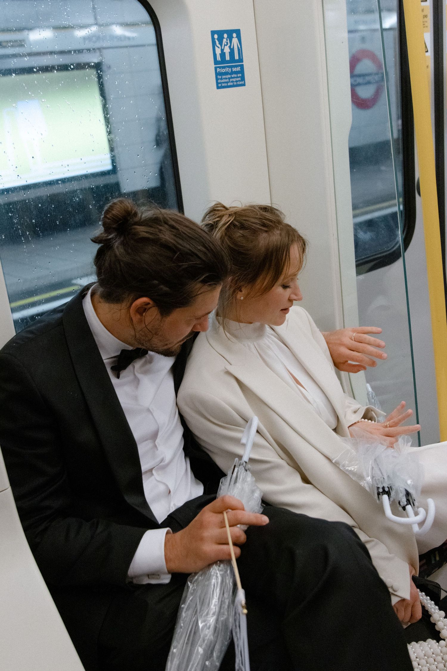 Newlywed couple in wedding attire riding London Underground tube train, bride in white suit and groom in black tuxedo.