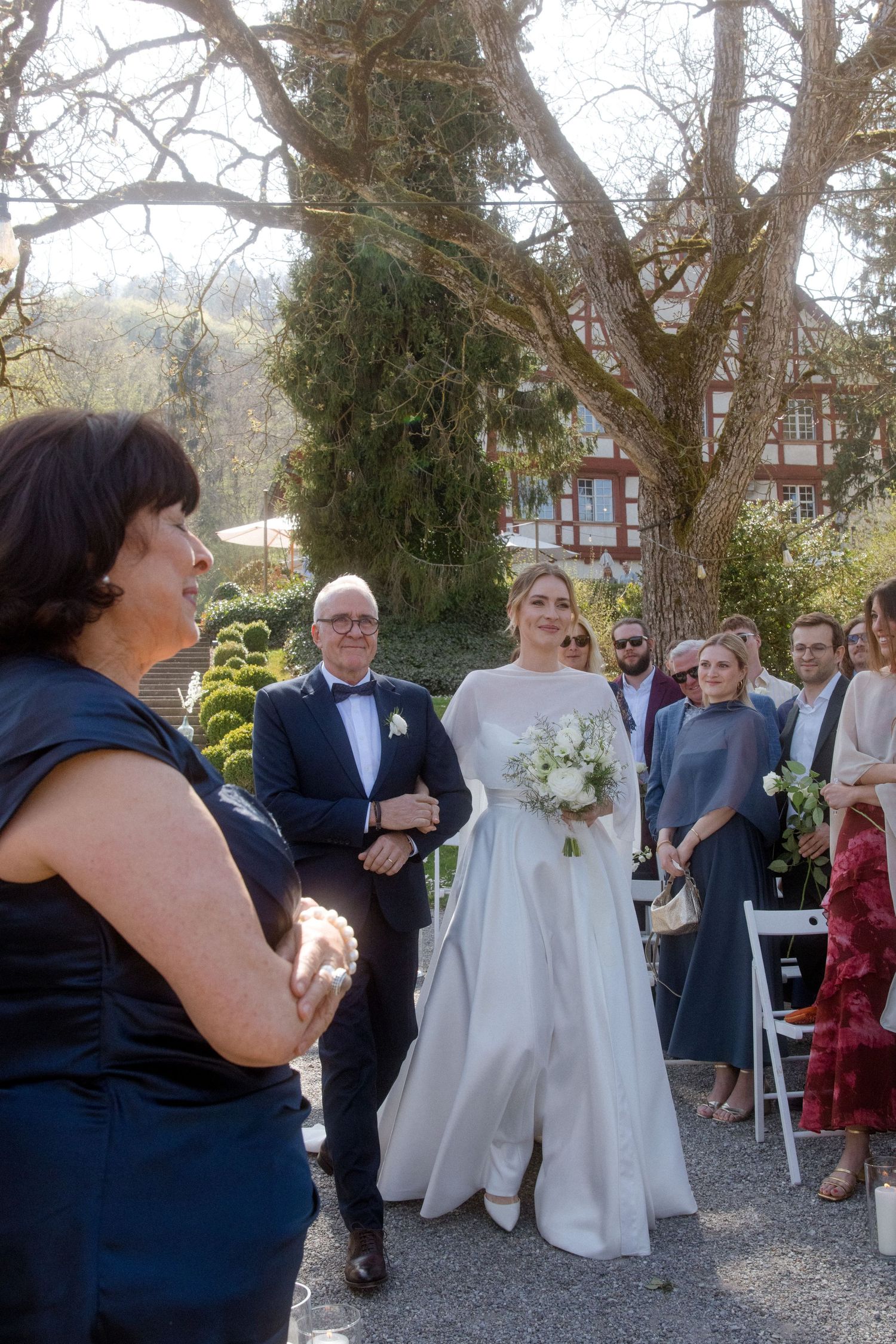 Bride in white gown holding bouquet walks toward officiant at outdoor wedding ceremony.