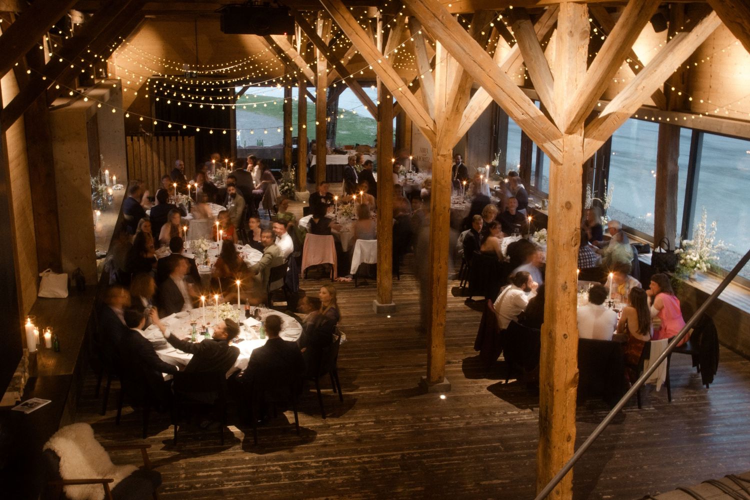 Elegant candlelit barn wedding reception with string lights, wooden beams, and guests dining at round tables.