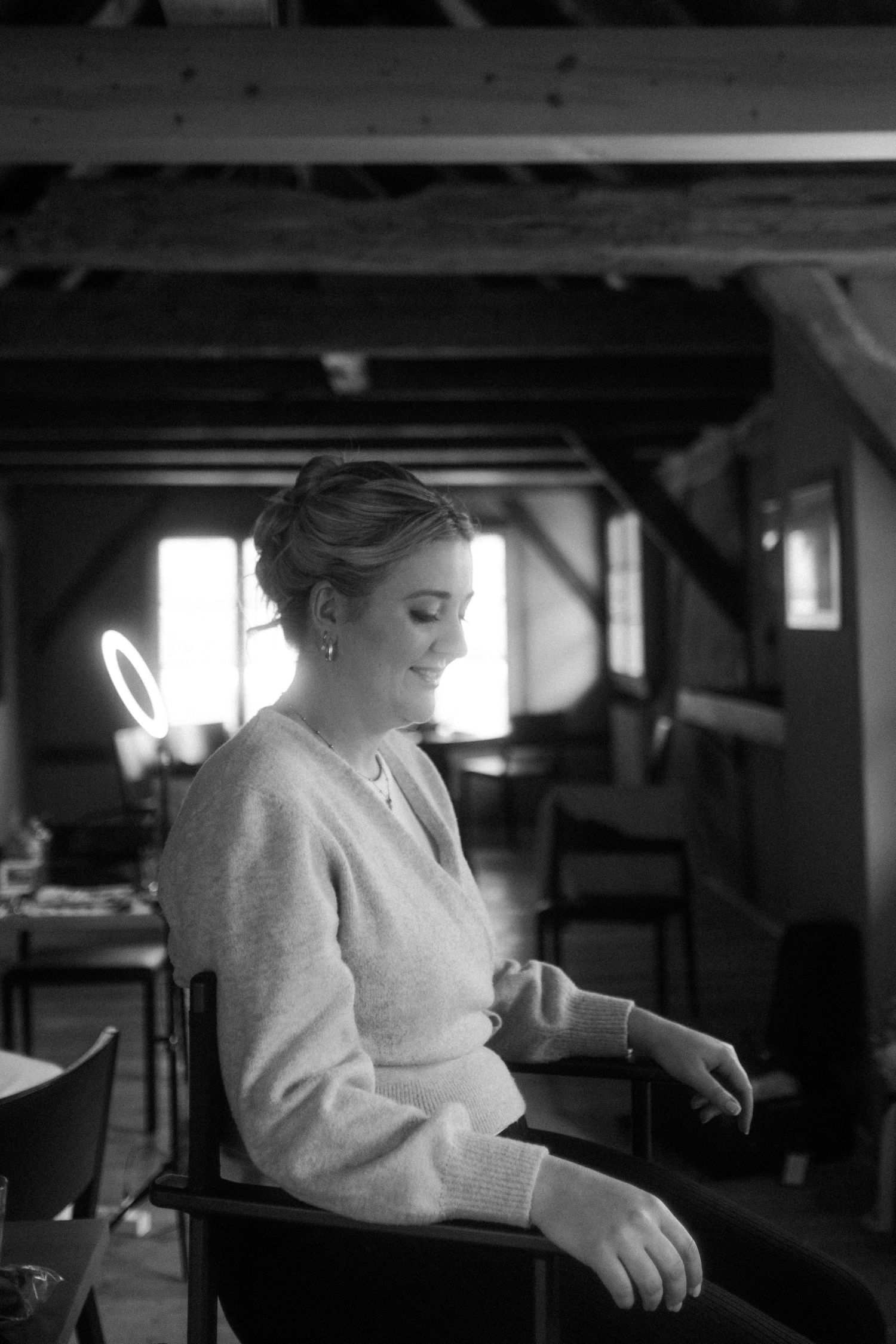 Black and white photo of a smiling woman in a cozy sweater seated in a rustic interior with wooden beams.