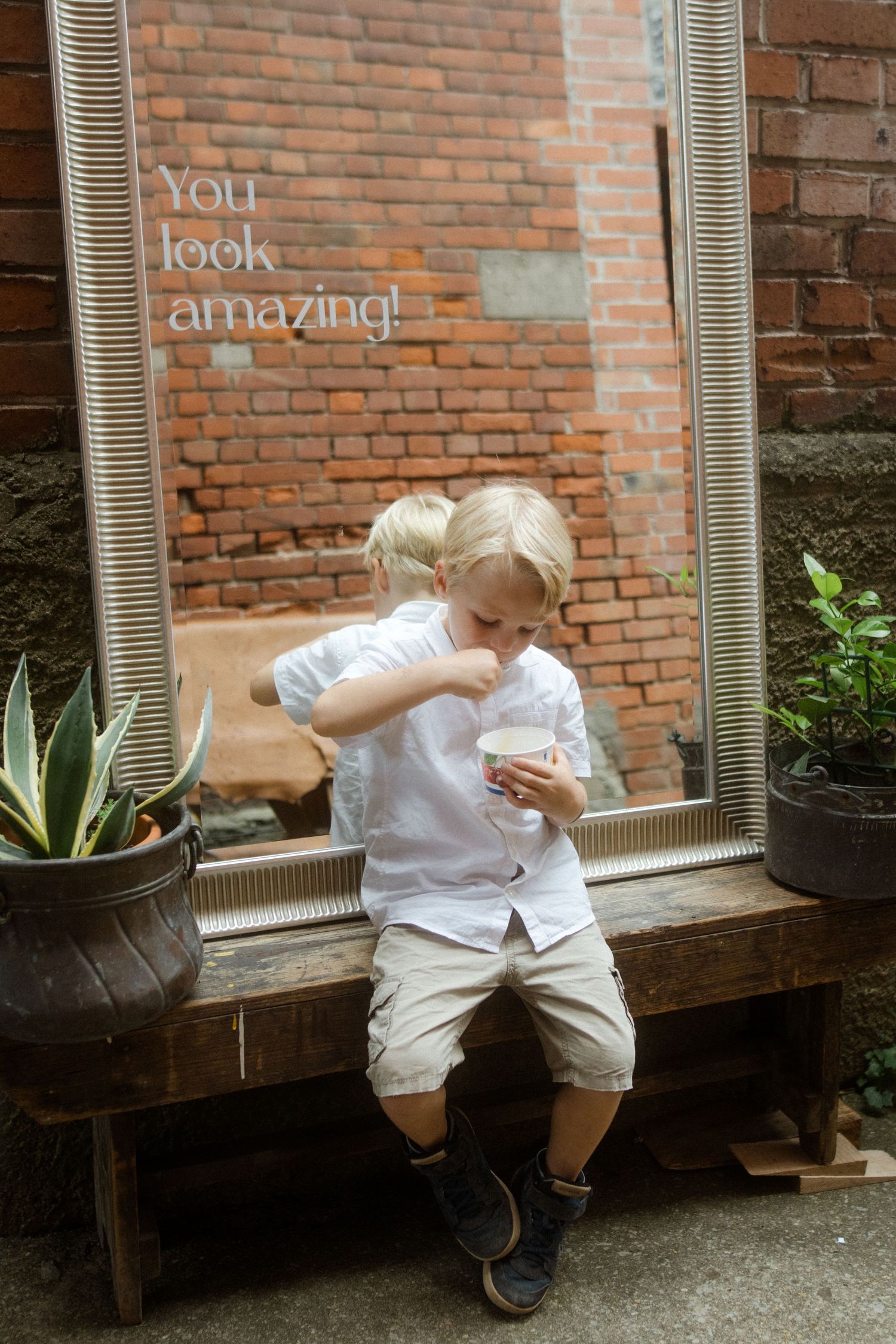 Young child in white outfit sits on wooden bench near brick wall.