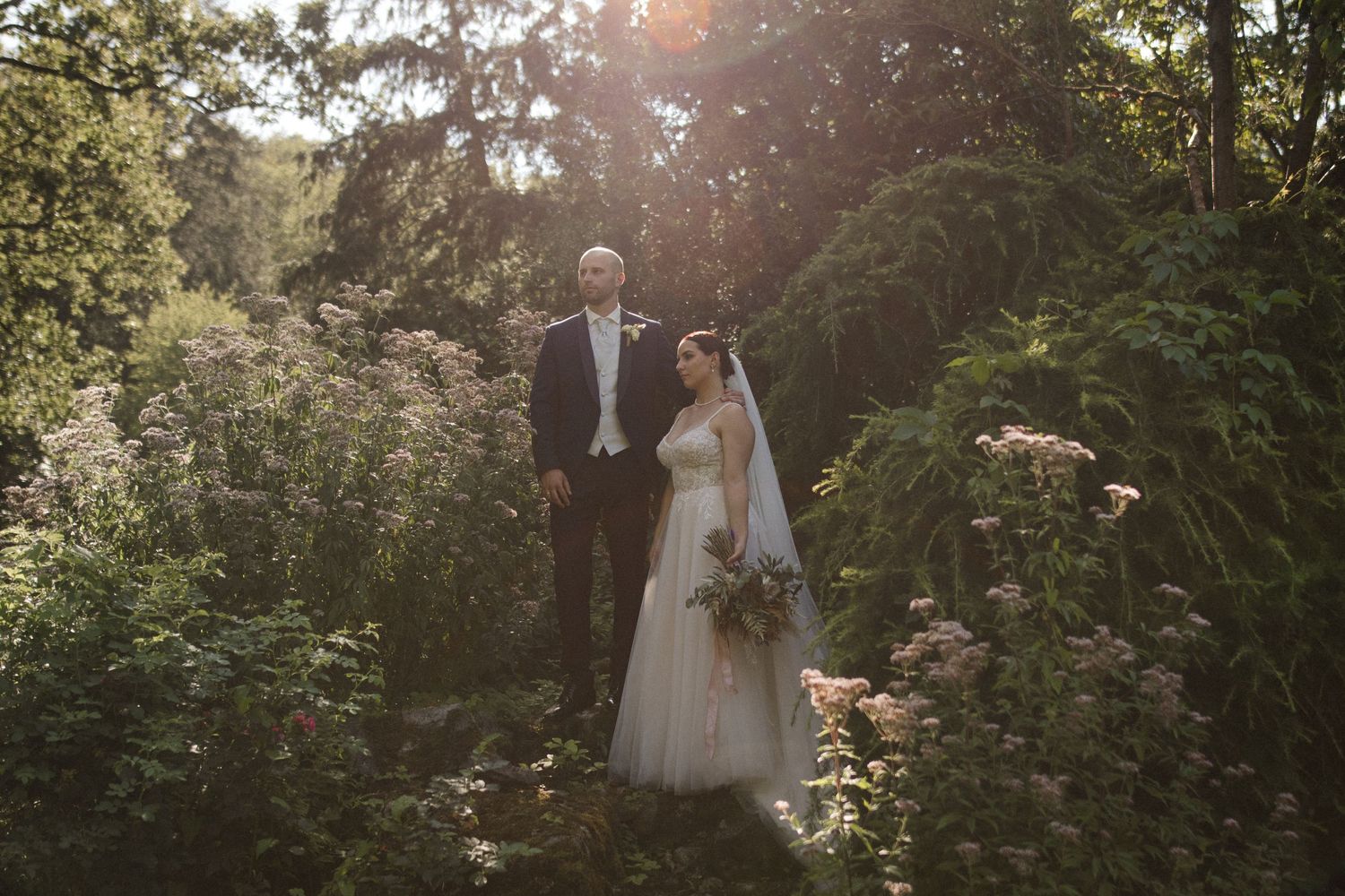 A bride and groom stand together in a lush garden, bathed in golden sunlight, on their wedding day.