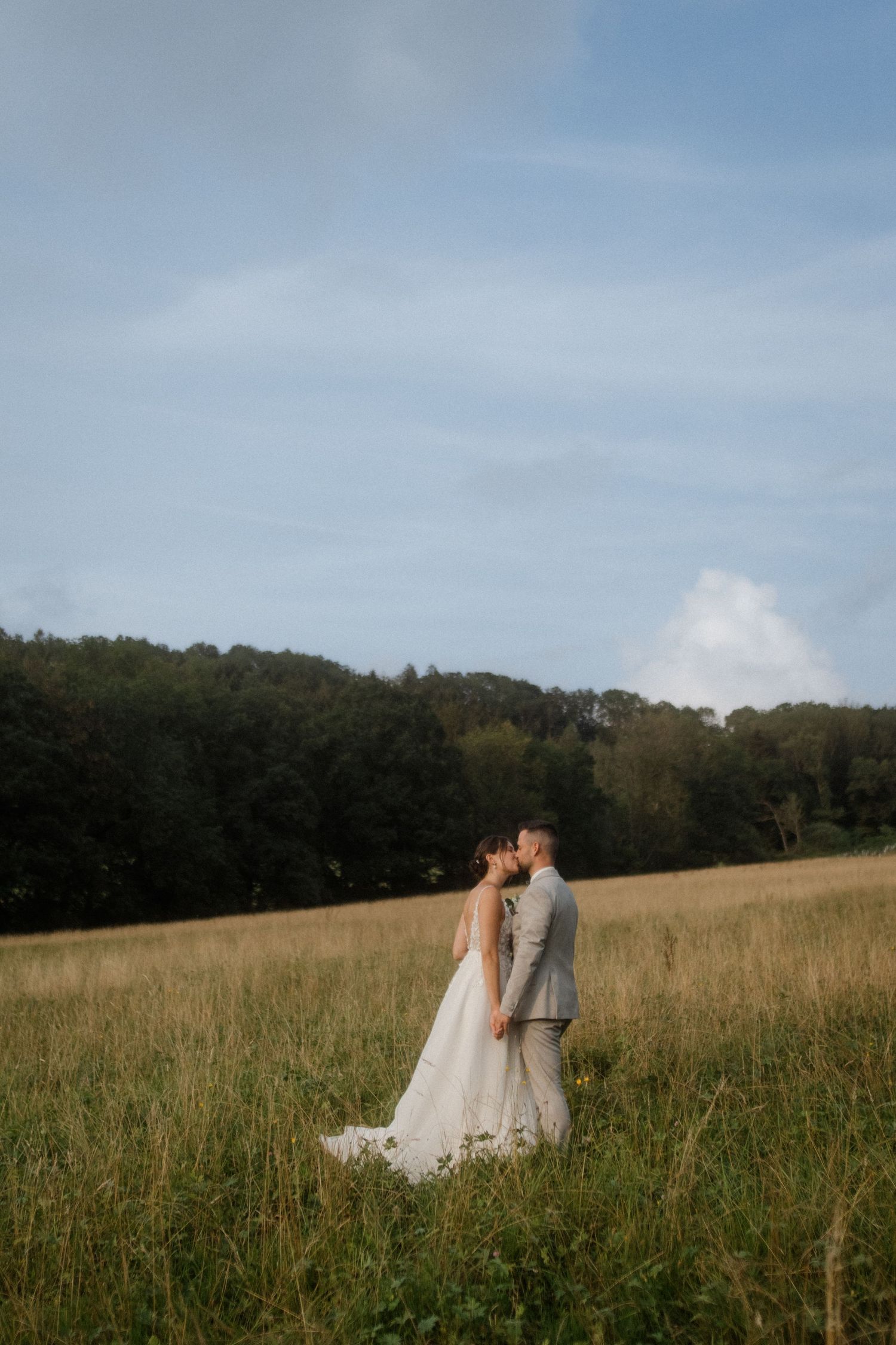 Two people in white dresses walking through a golden meadow with trees in the background.