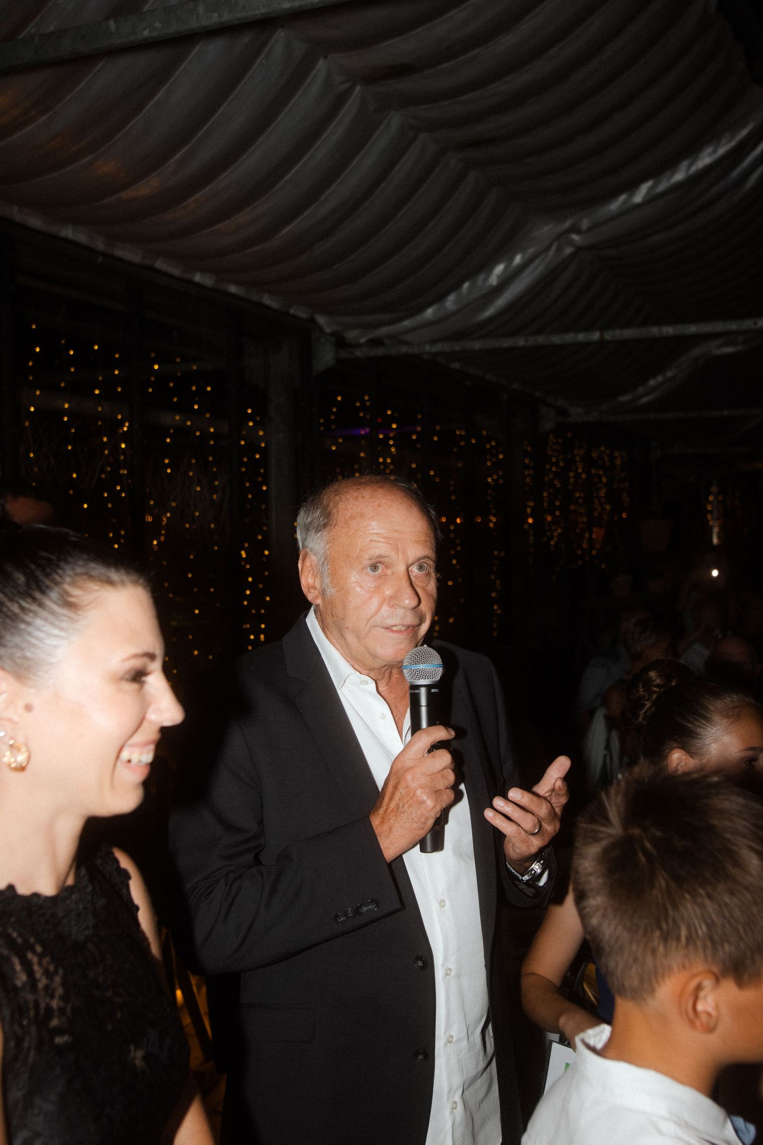 Speaker holds microphone at wedding reception with guests gathered around.
