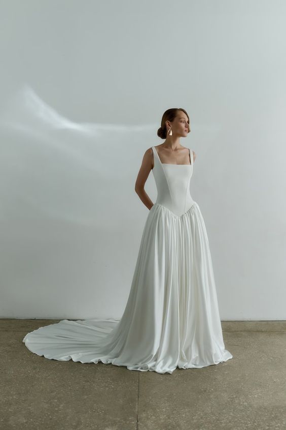 Minimalist white wedding gown with pleated skirt and elegant train against grey backdrop.