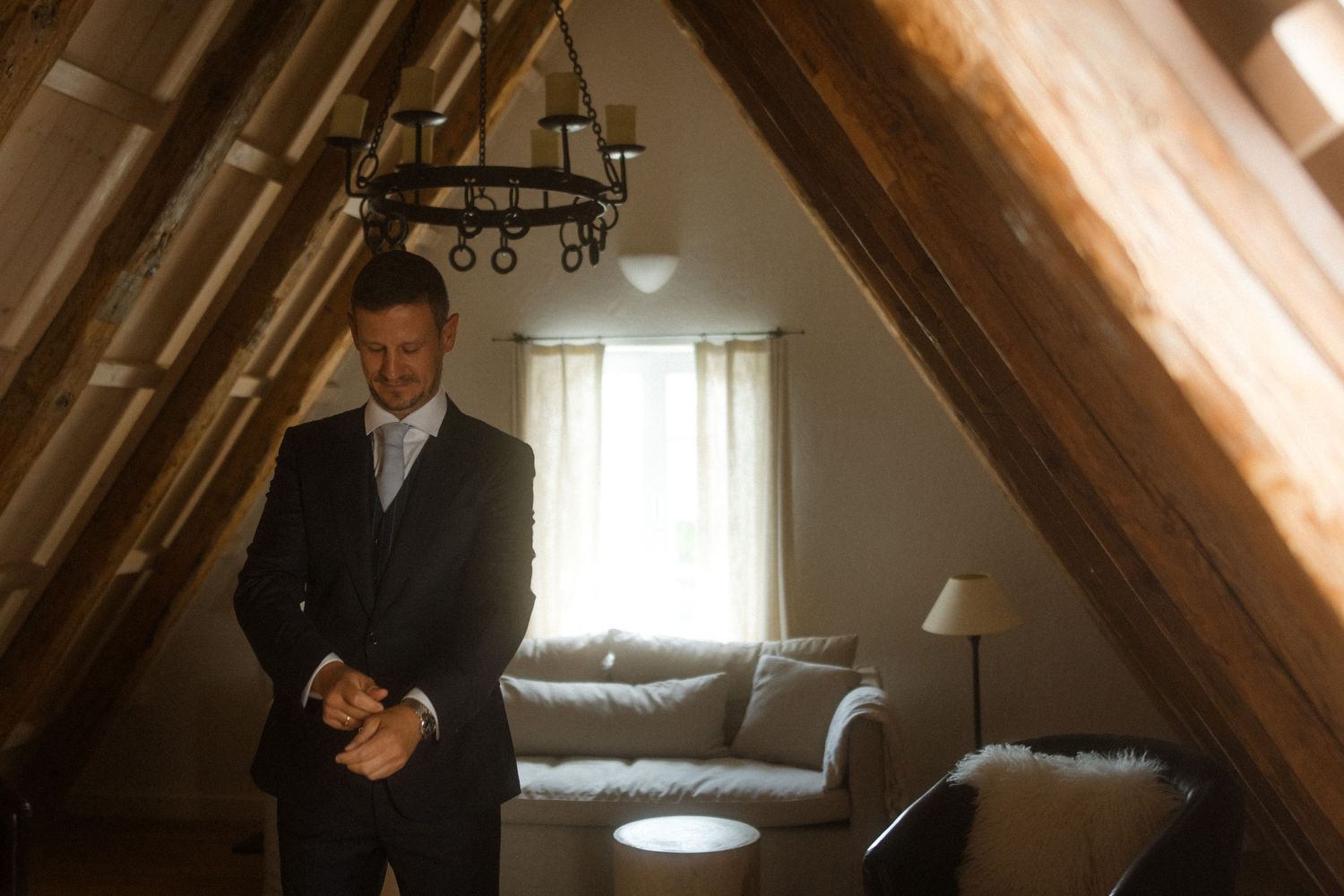 Well-dressed man standing in an attic room with exposed wooden beams and leather furniture.