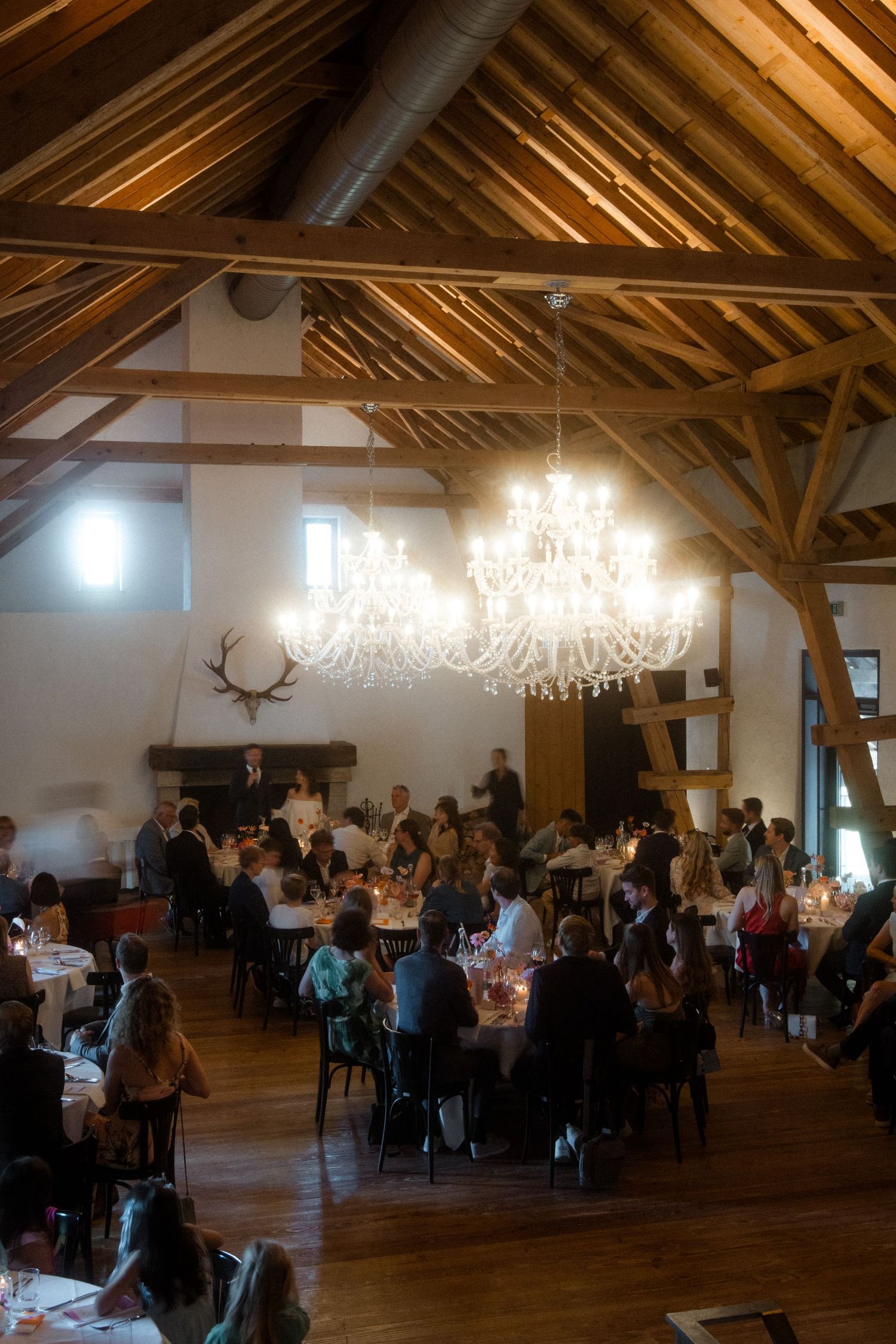 Elegant wedding reception in rustic venue with exposed wooden beams and crystal chandeliers illuminating dining tables.