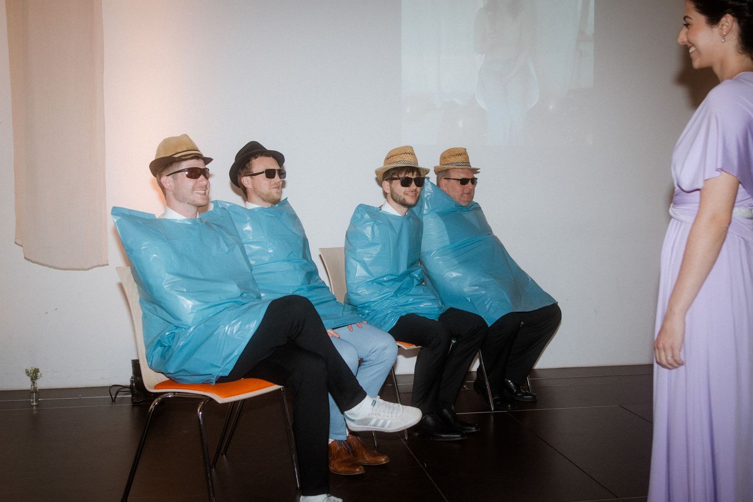 Four people in blue ponchos and hats sit in chairs watching a projected screen.