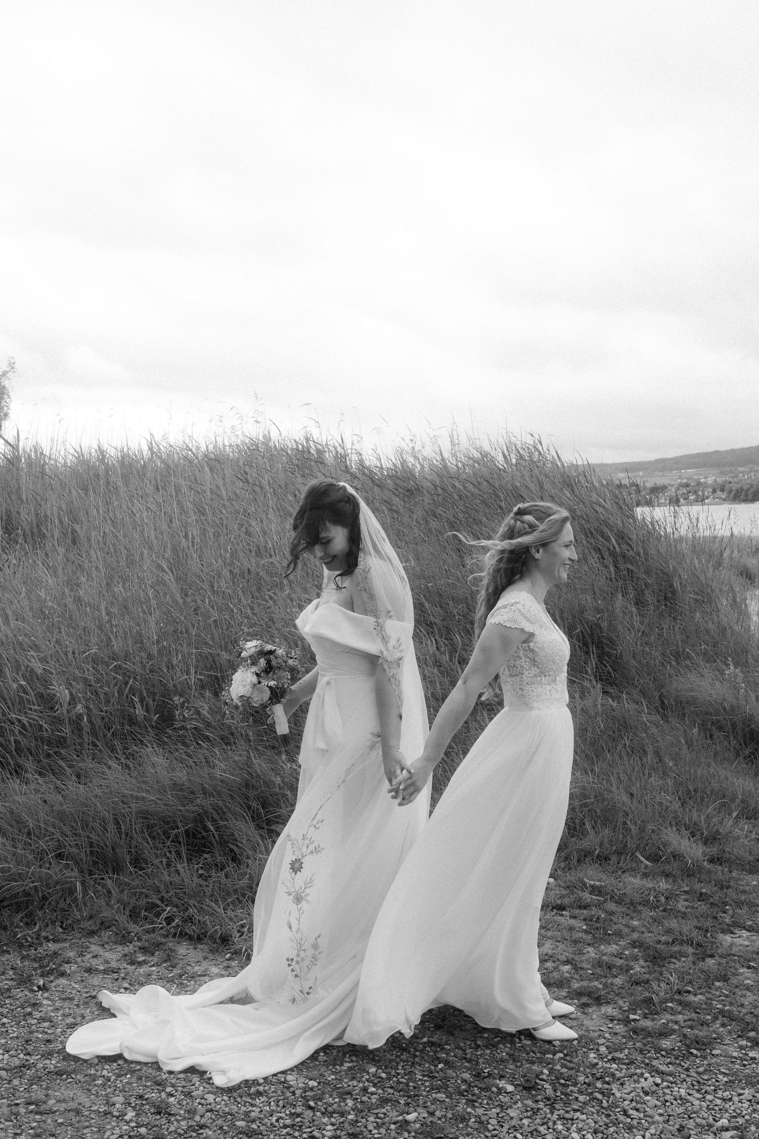 Two figures in white wedding dresses pose dramatically in a windswept field of tall grass.