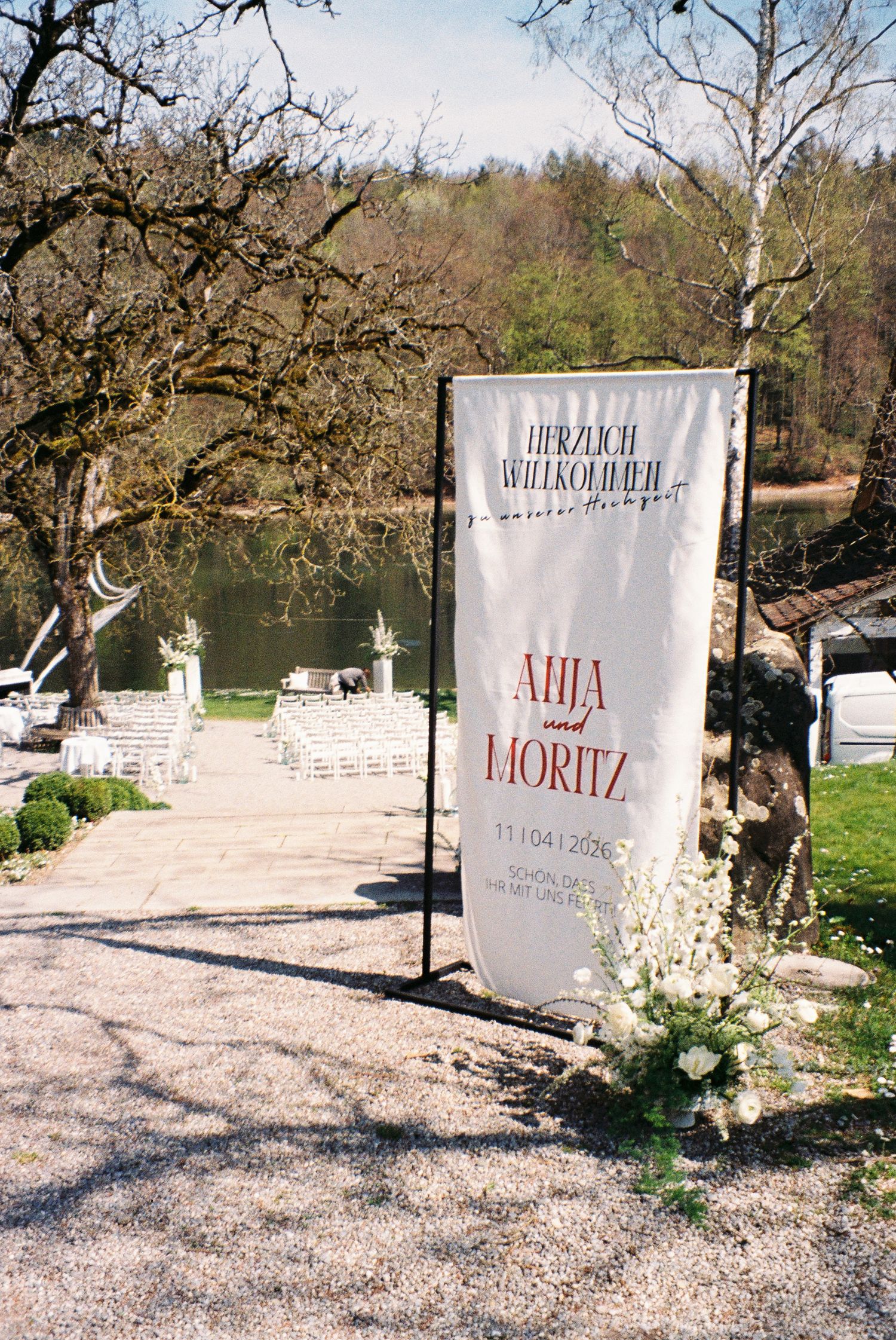 Outdoor wedding ceremony setup with welcome banner for Ahja and Moritz, dated 11/04/2026, near a scenic lake.