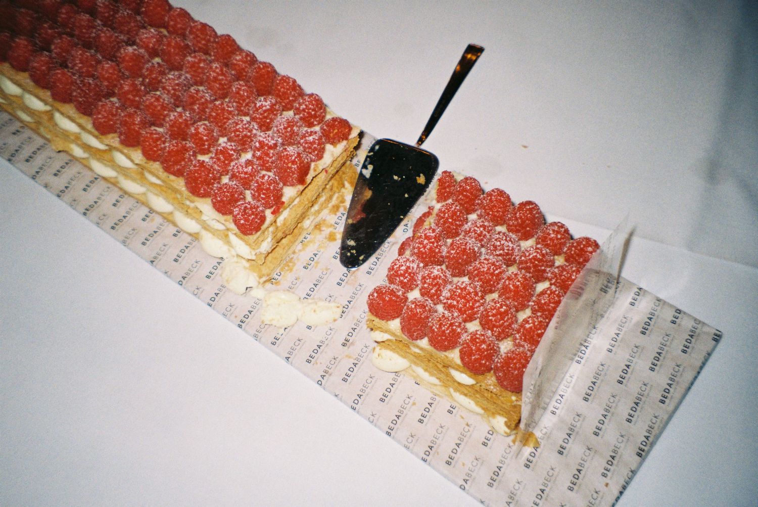 Elegant raspberry mille-feuille pastry cake with cream layers, topped with fresh raspberries, partially sliced on decorative paper.