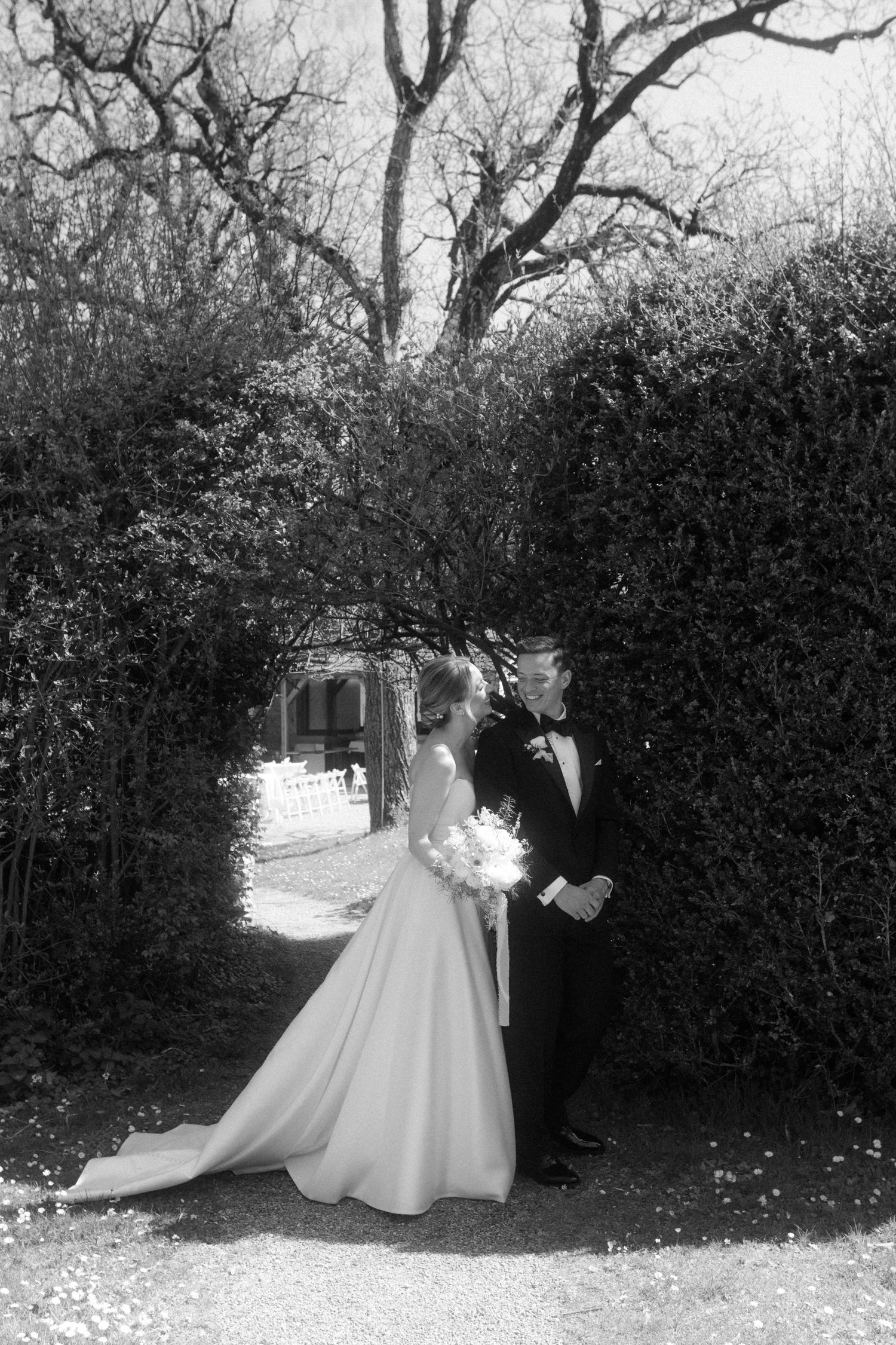 Black and white photo of bride and groom standing in a garden archway, bride holding bouquet.