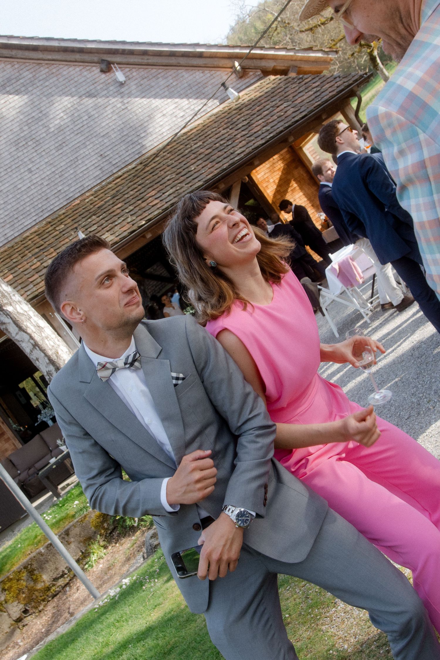 Couple dancing joyfully at outdoor wedding reception, man in gray suit with bow tie lifting woman in pink jumpsuit.