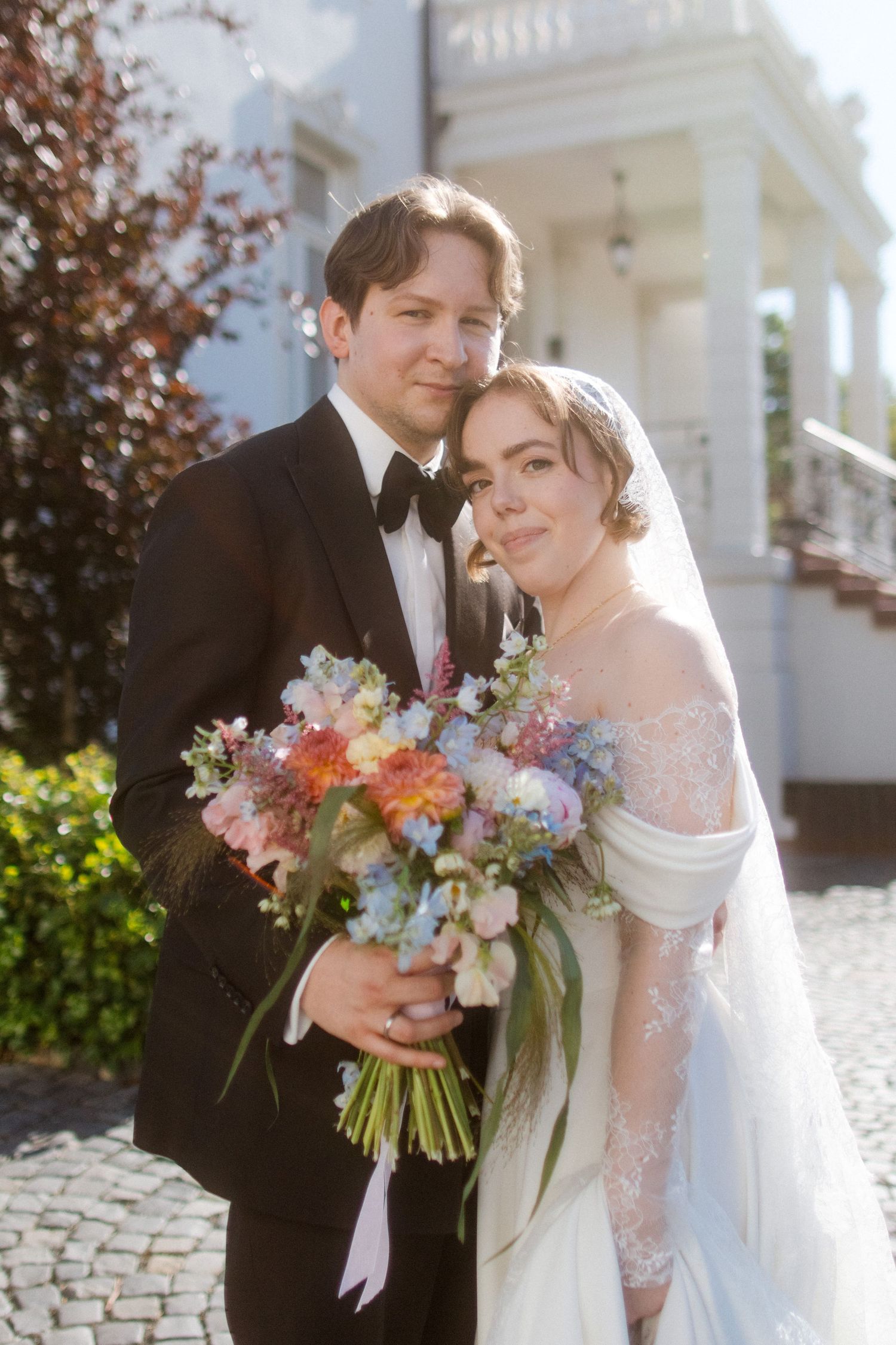 Bride and groom pose outdoors holding colorful bouquet, groom in tuxedo, bride in lace off-shoulder gown with veil.