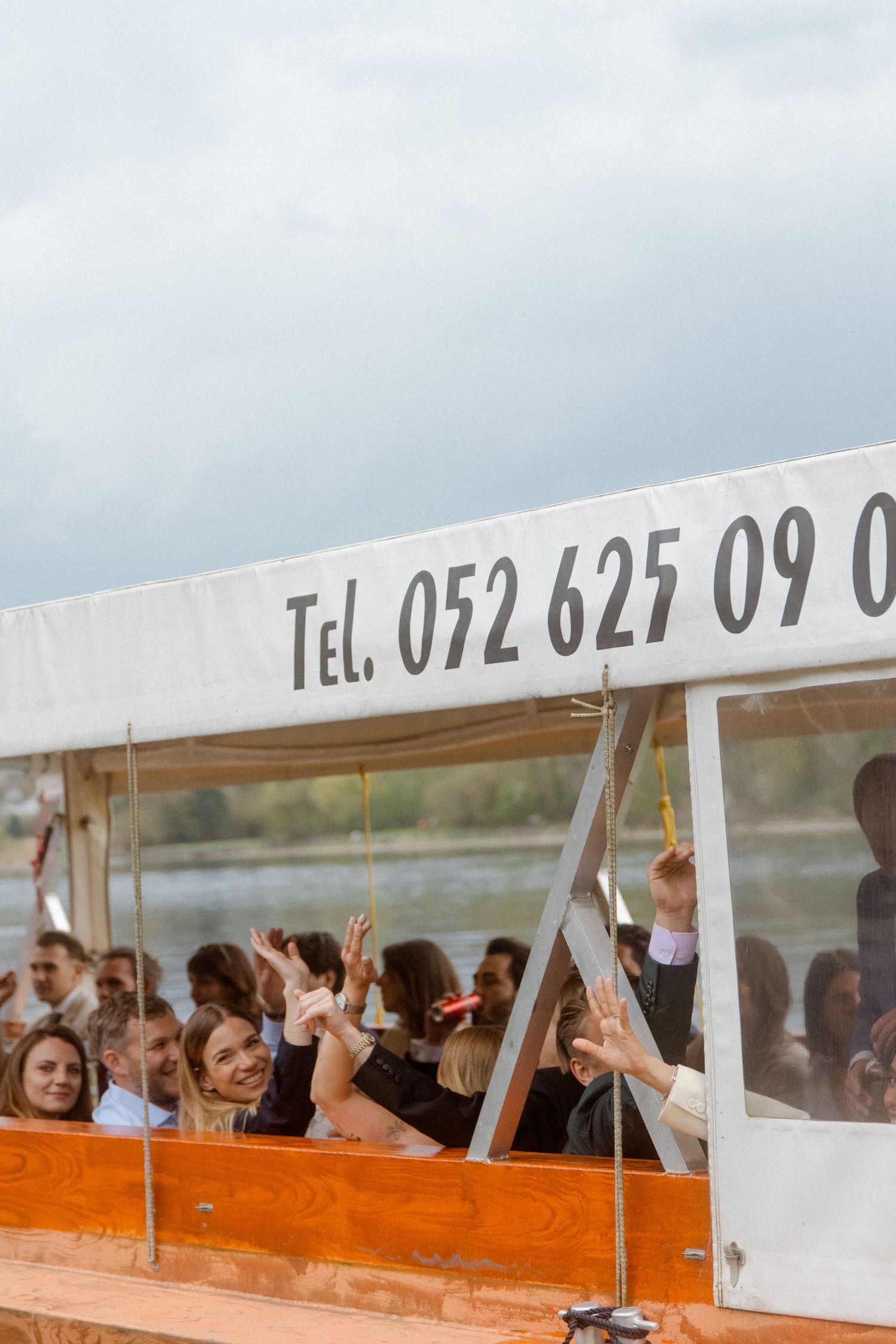 Wedding guests waving from inside covered boat on cloudy day on water.