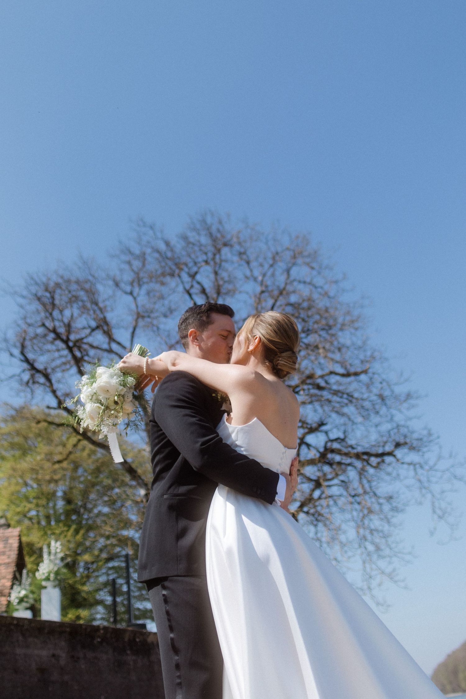 Bride and groom sharing a romantic kiss outdoors on their wedding day, with a clear blue sky and bare trees behind them.