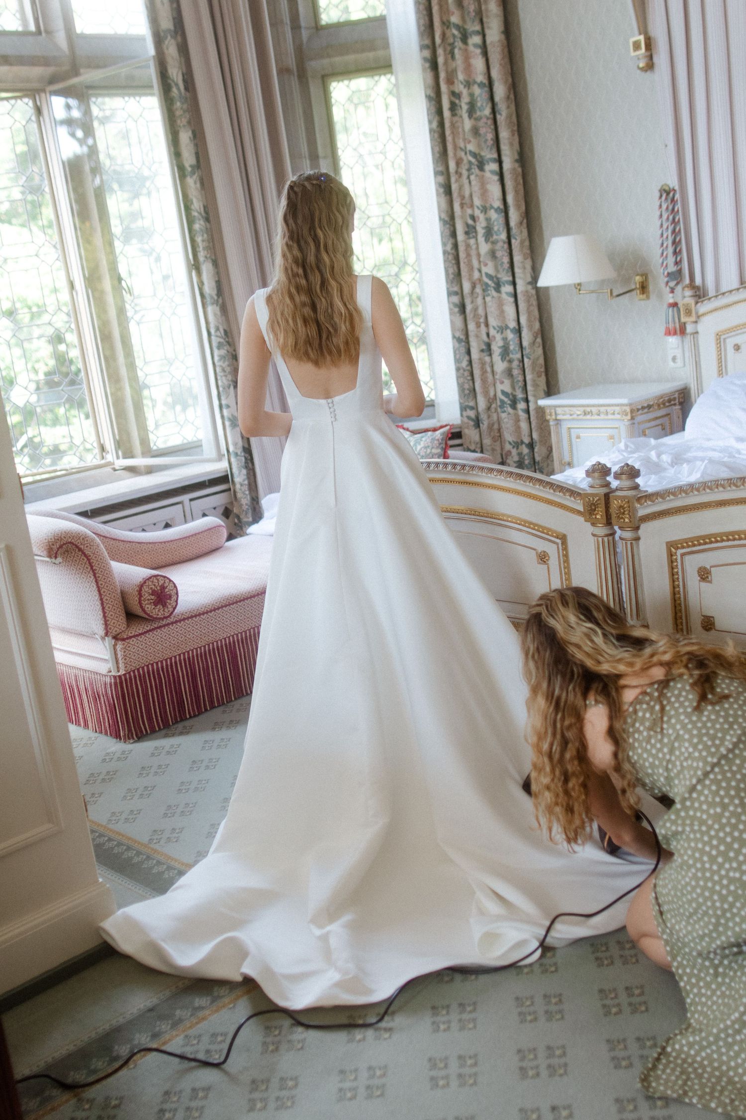Person in white gown moves through an elegant room with vintage furniture and floral curtains.