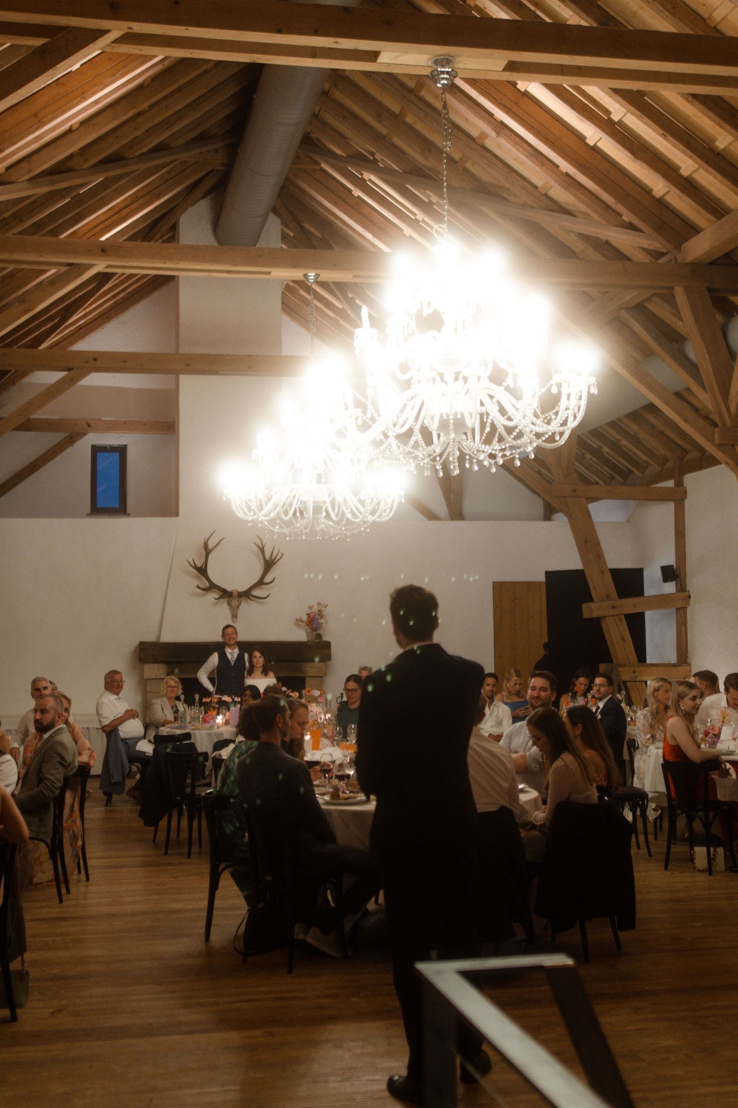 Rustic venue with exposed wooden beams, chandeliers, and guests seated at dinner tables.