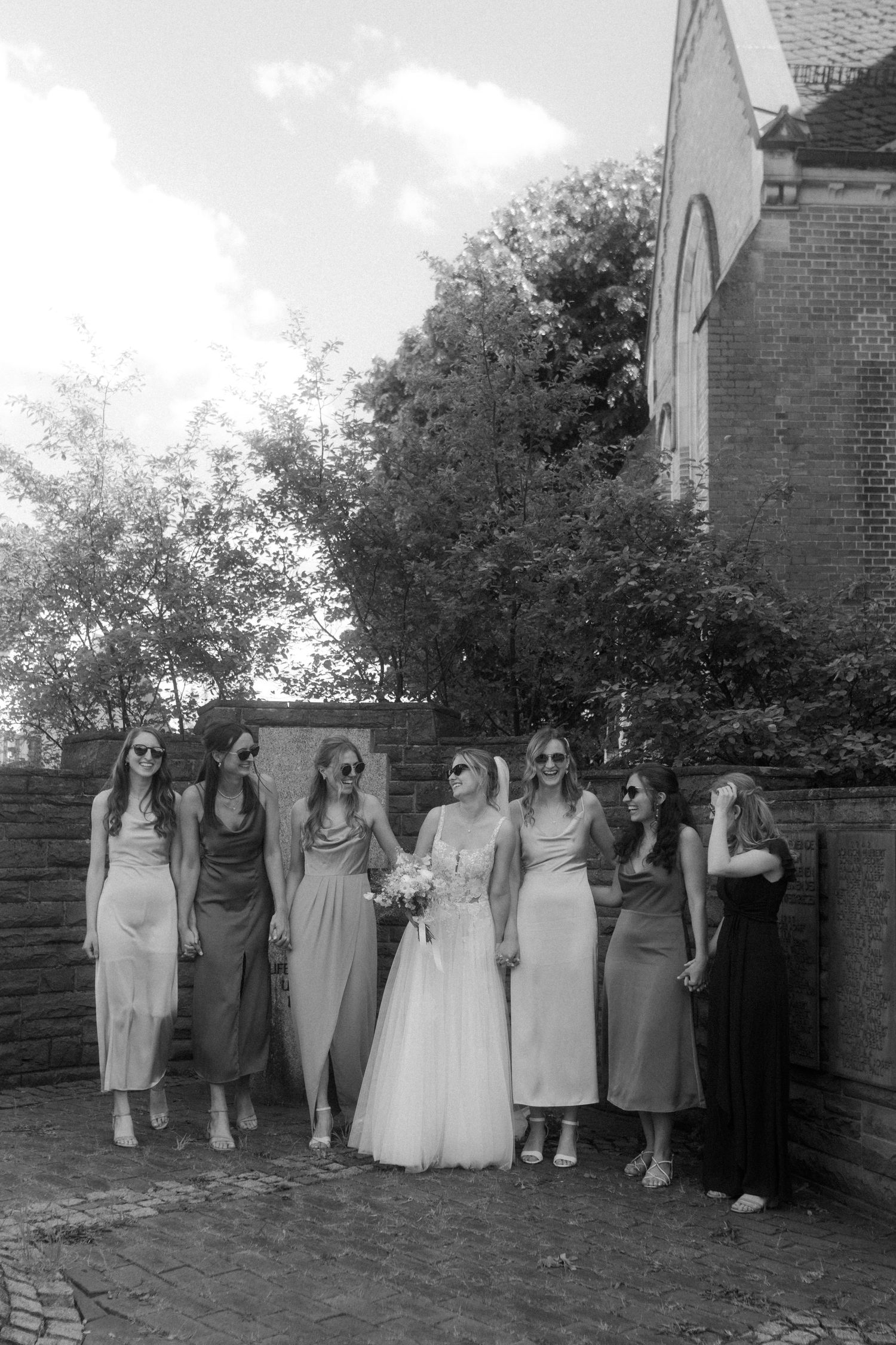 Black and white photo of bridesmaids in elegant dresses posing with bride outside church.
