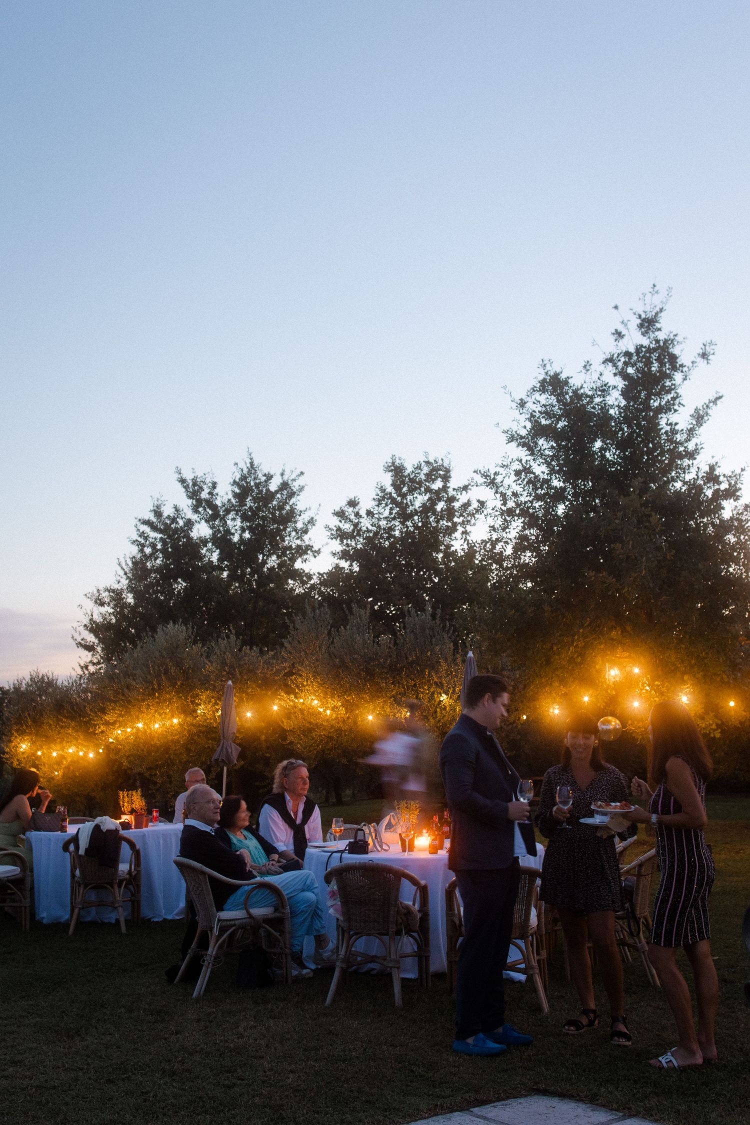 An outdoor evening party setup with string lights illuminating tables and trees.