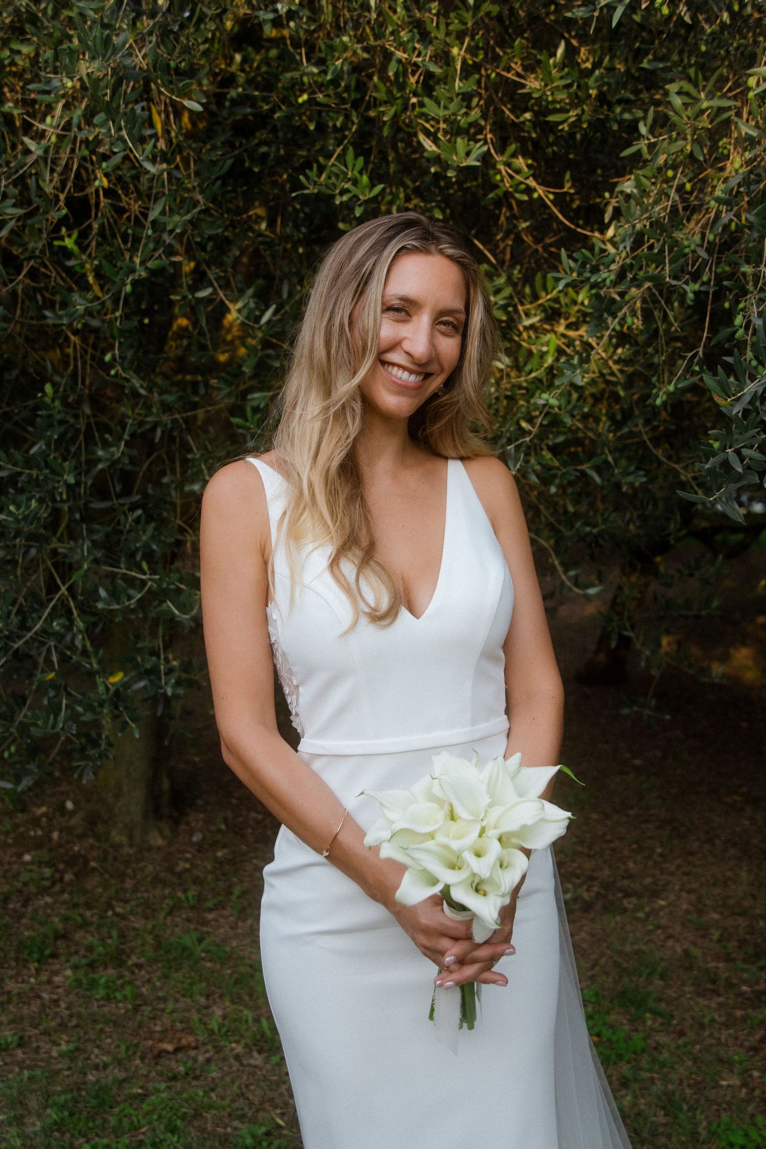 Elegant bride in a fitted white dress holding a bouquet of white flowers in a garden setting.