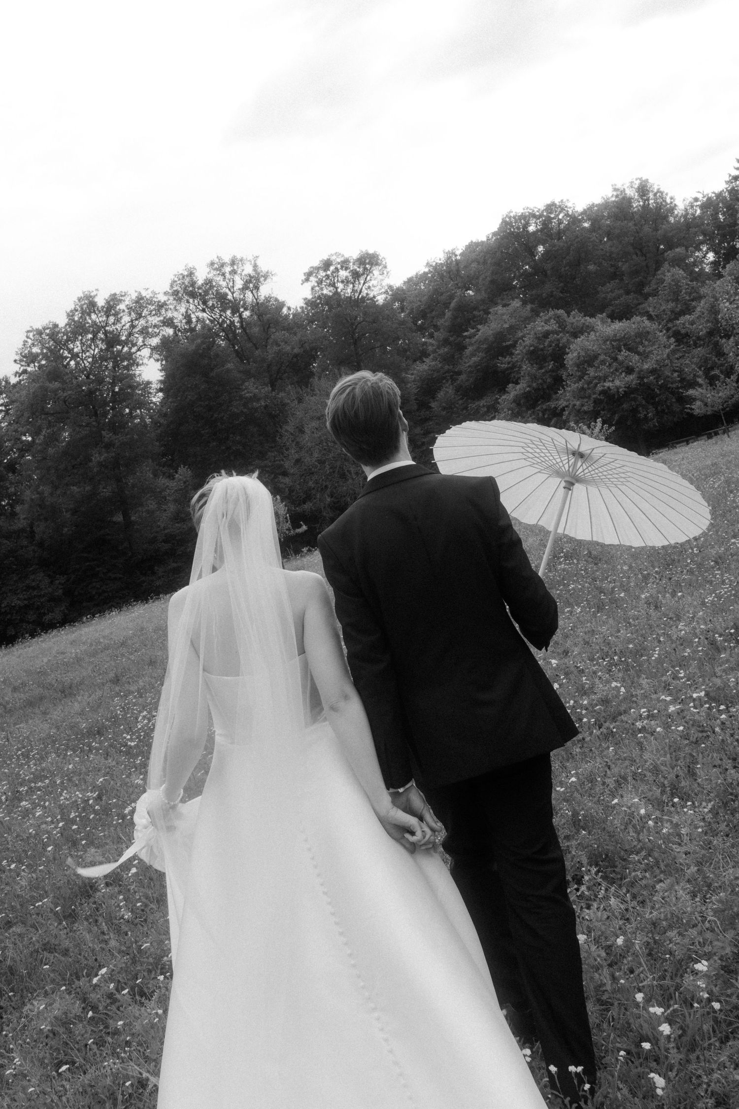 A couple walks through a wildflower meadow holding a white parasol.