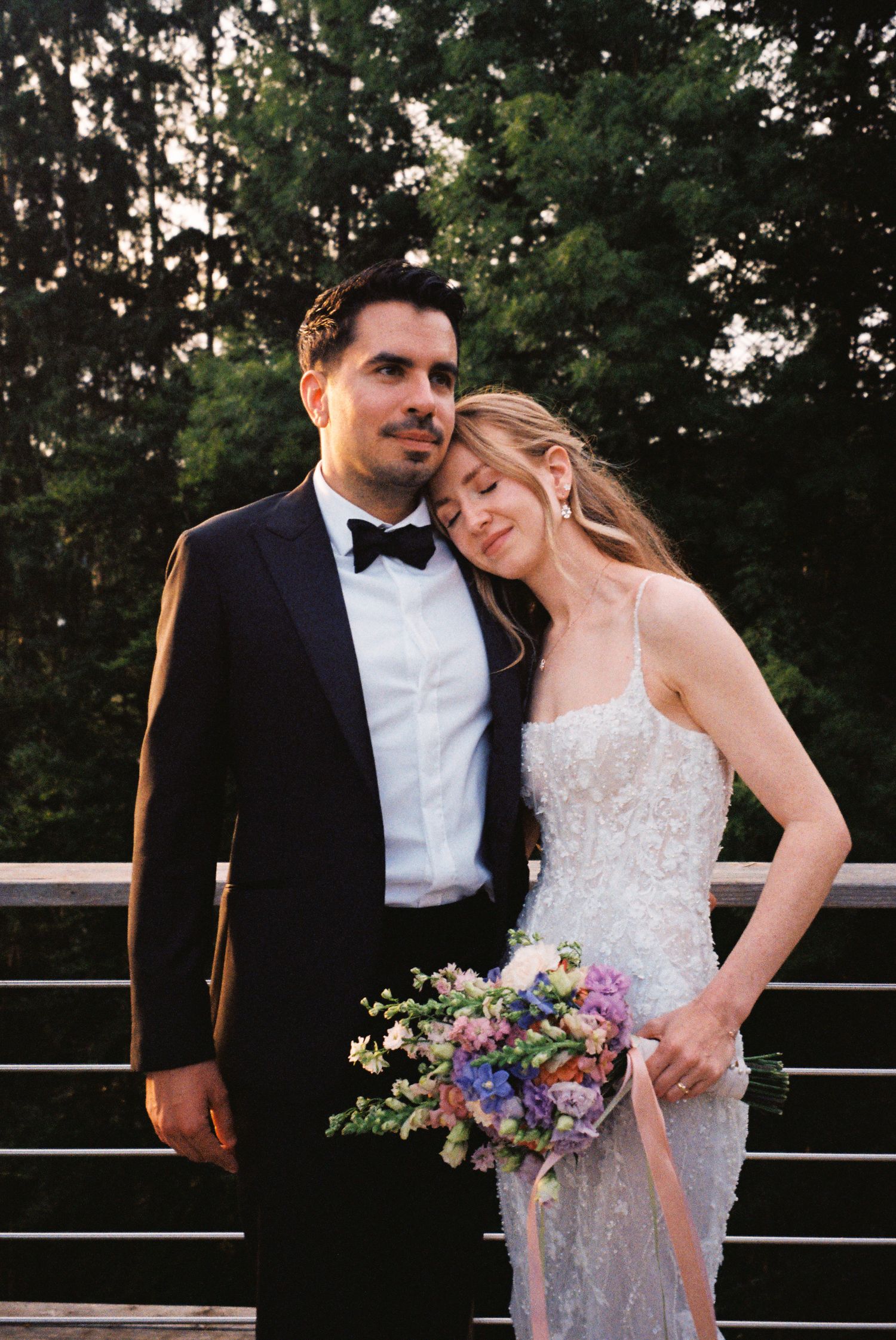 A couple in formal wedding attire stands on a deck at sunset with a colorful bouquet of flowers.