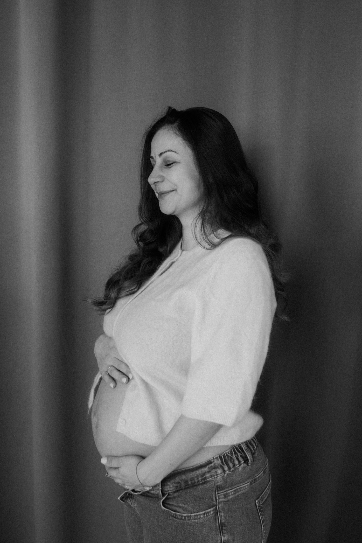 A black and white portrait of an expectant mother in casual attire smiling while holding her belly.