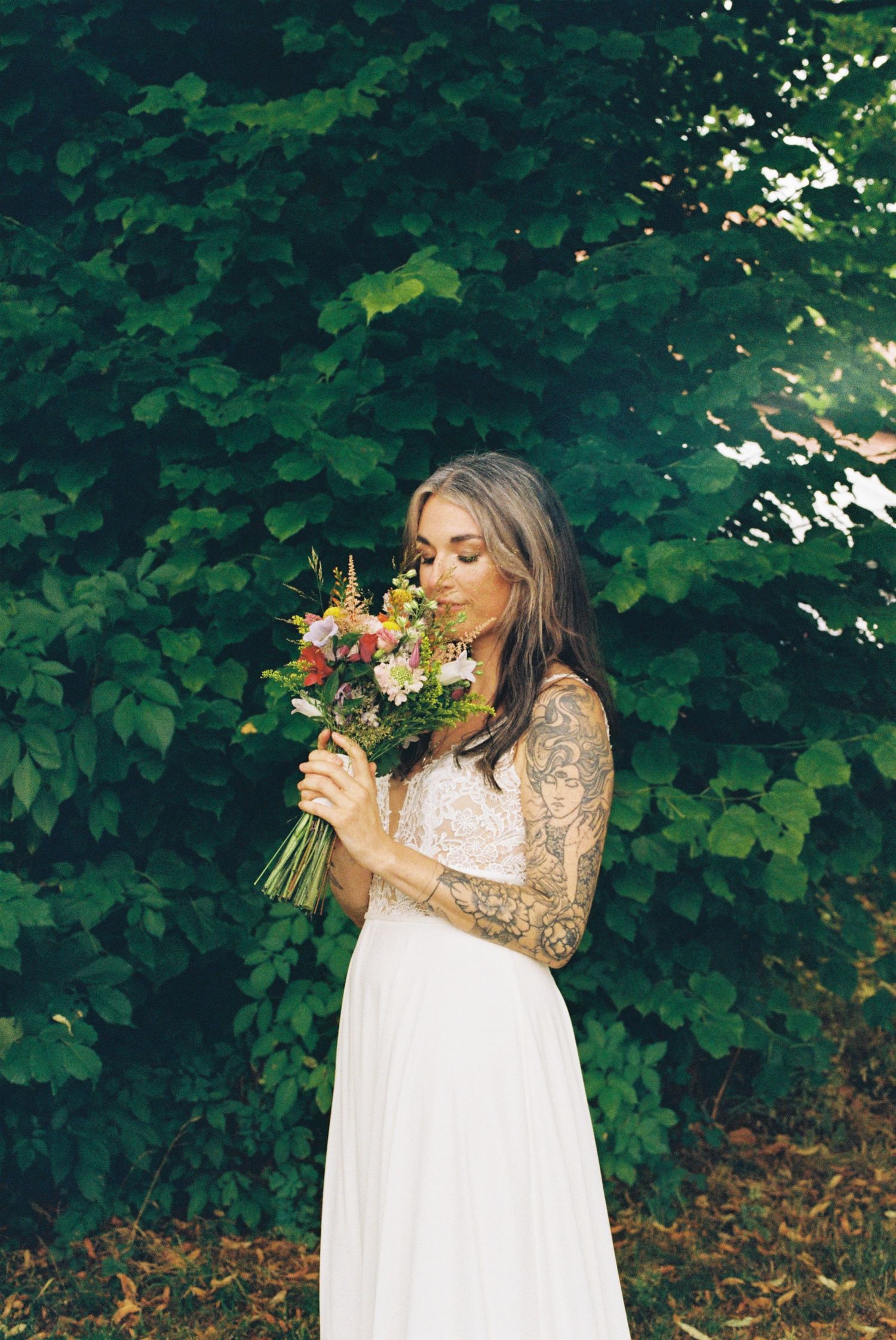 A bride in a white dress with tattoo sleeves smells a colorful bouquet against a backdrop of green foliage.