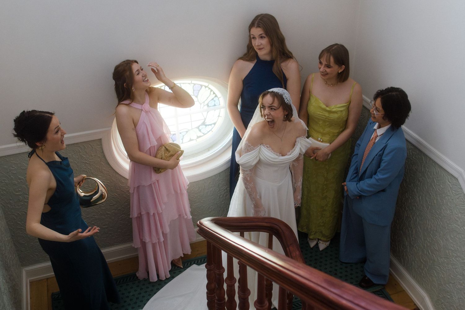 Bride in white gown with bridesmaids in colorful dresses gathered on elegant staircase near stained glass window.