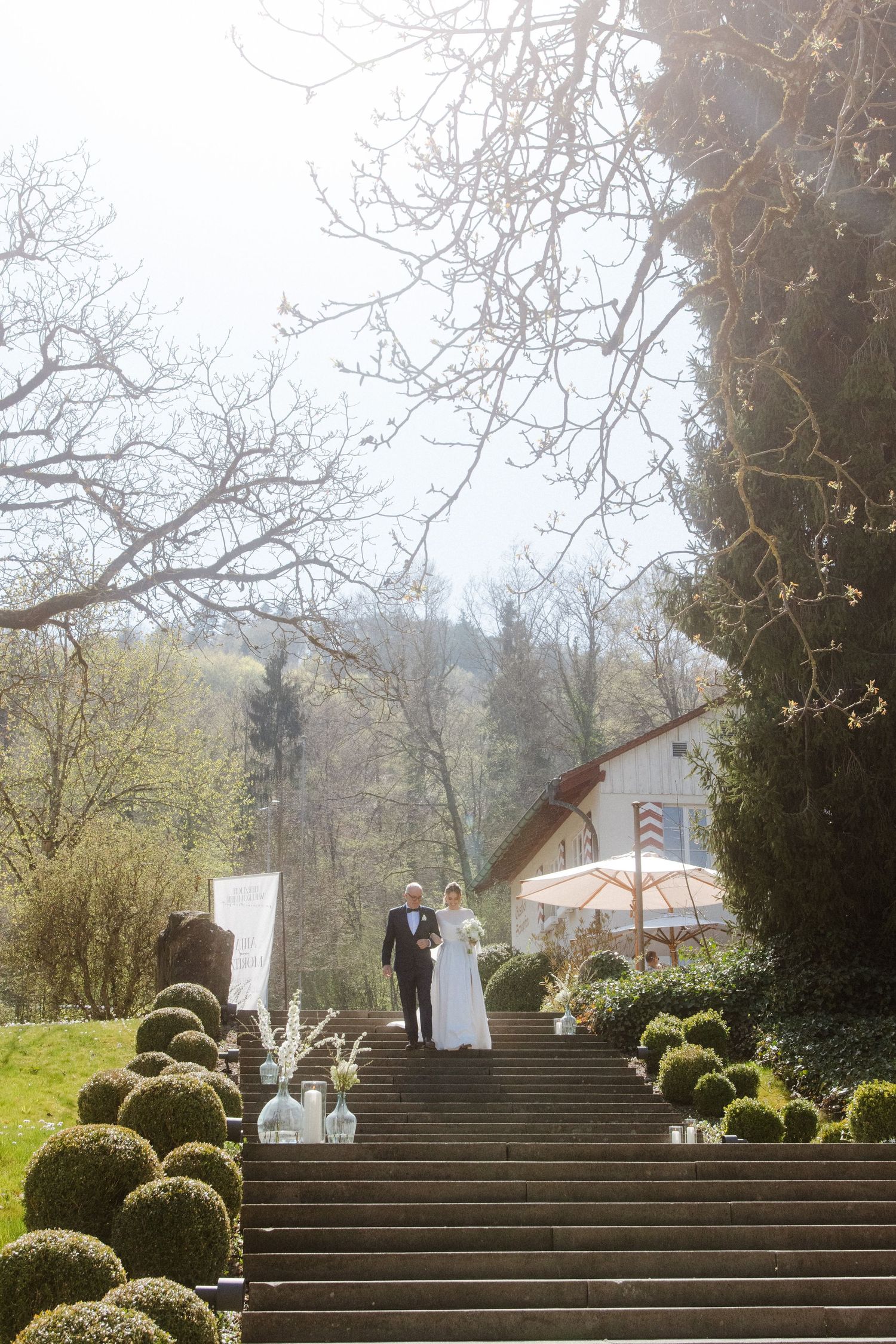 Bride and groom descend elegant stone stairs lined with topiary bushes at an outdoor wedding venue.