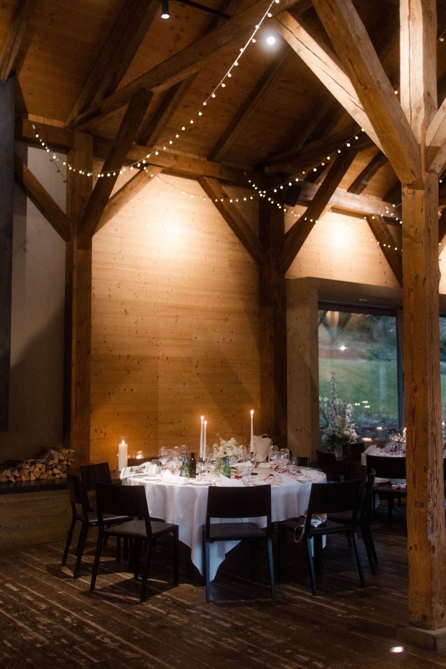 Rustic barn wedding reception with candlelit round tables, string lights, and wooden beam architecture.