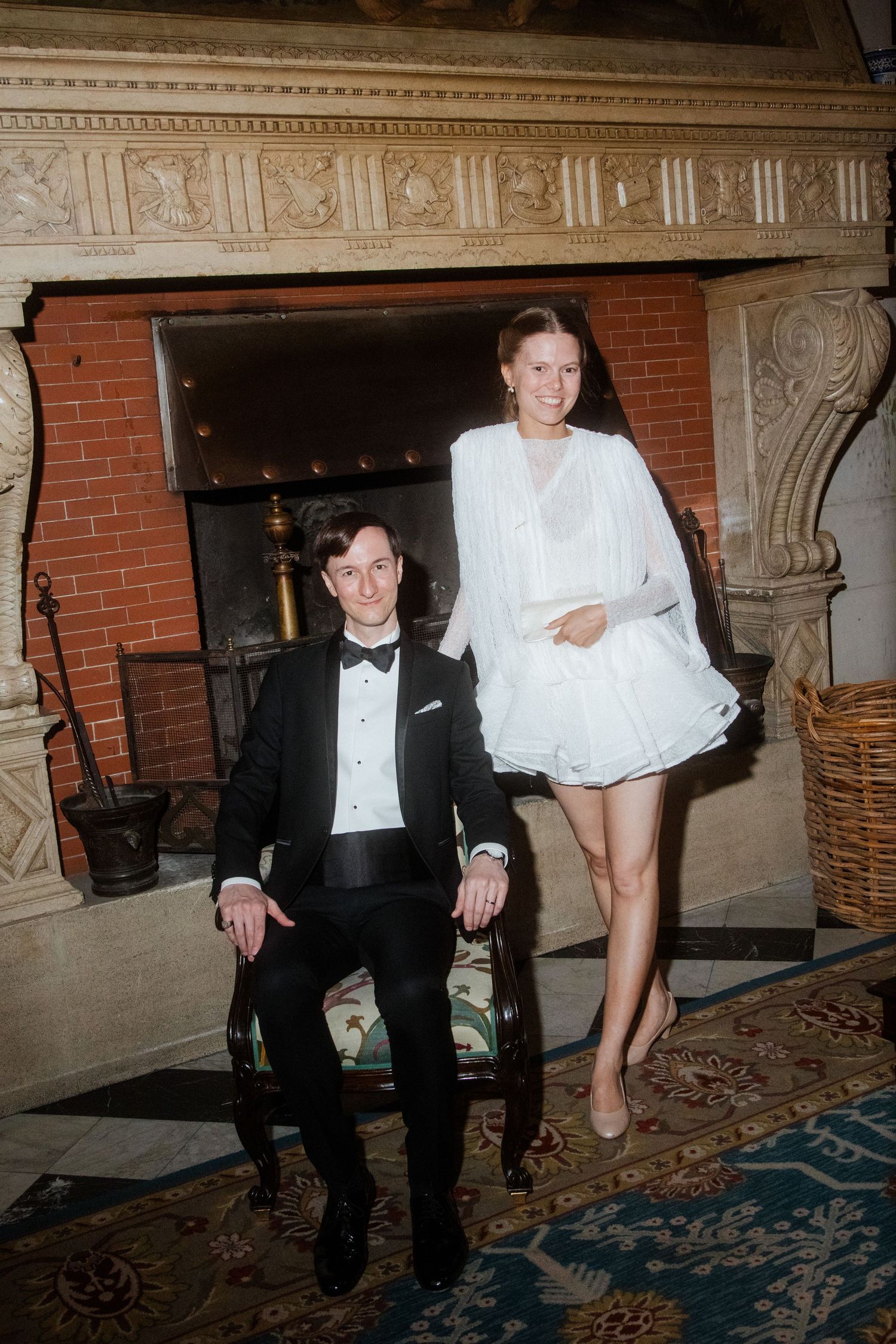 Couple in formal attire relaxing by ornate brick fireplace on patterned carpet.